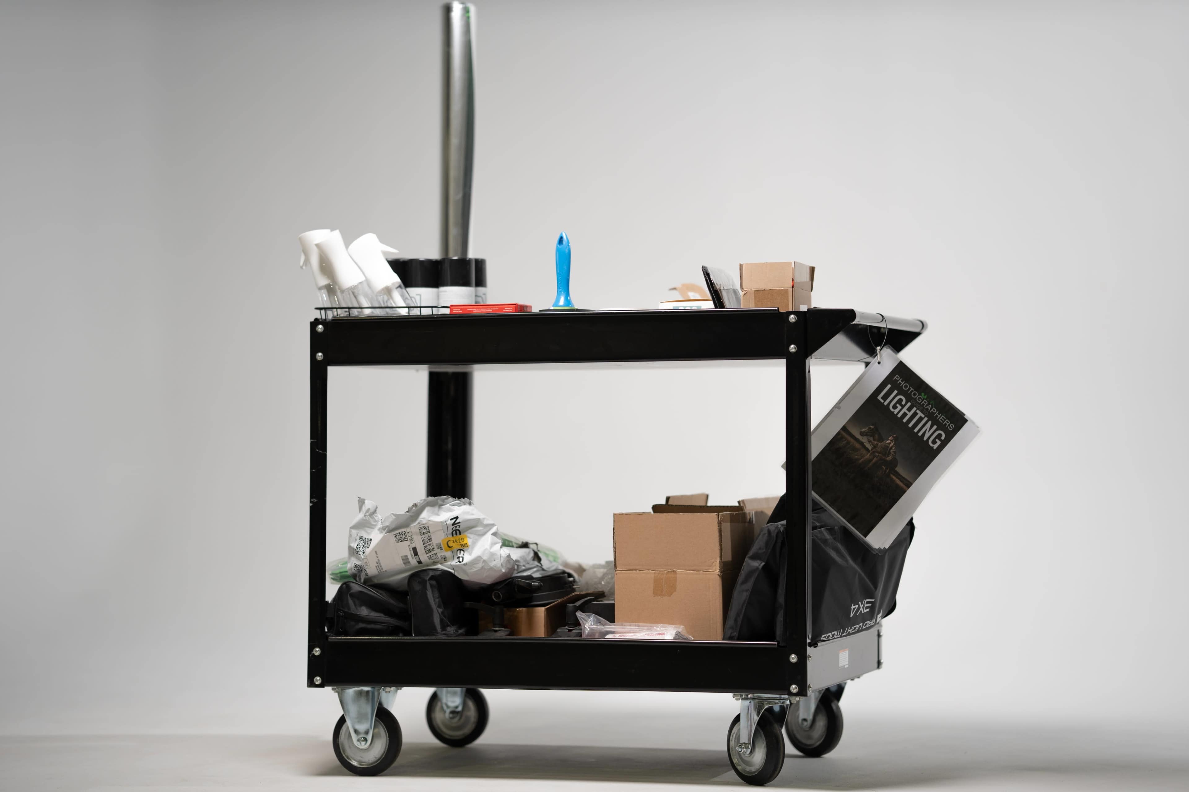 A black utility cart with various items including tools, boxes, and a stack of papers is positioned on a smooth surface.