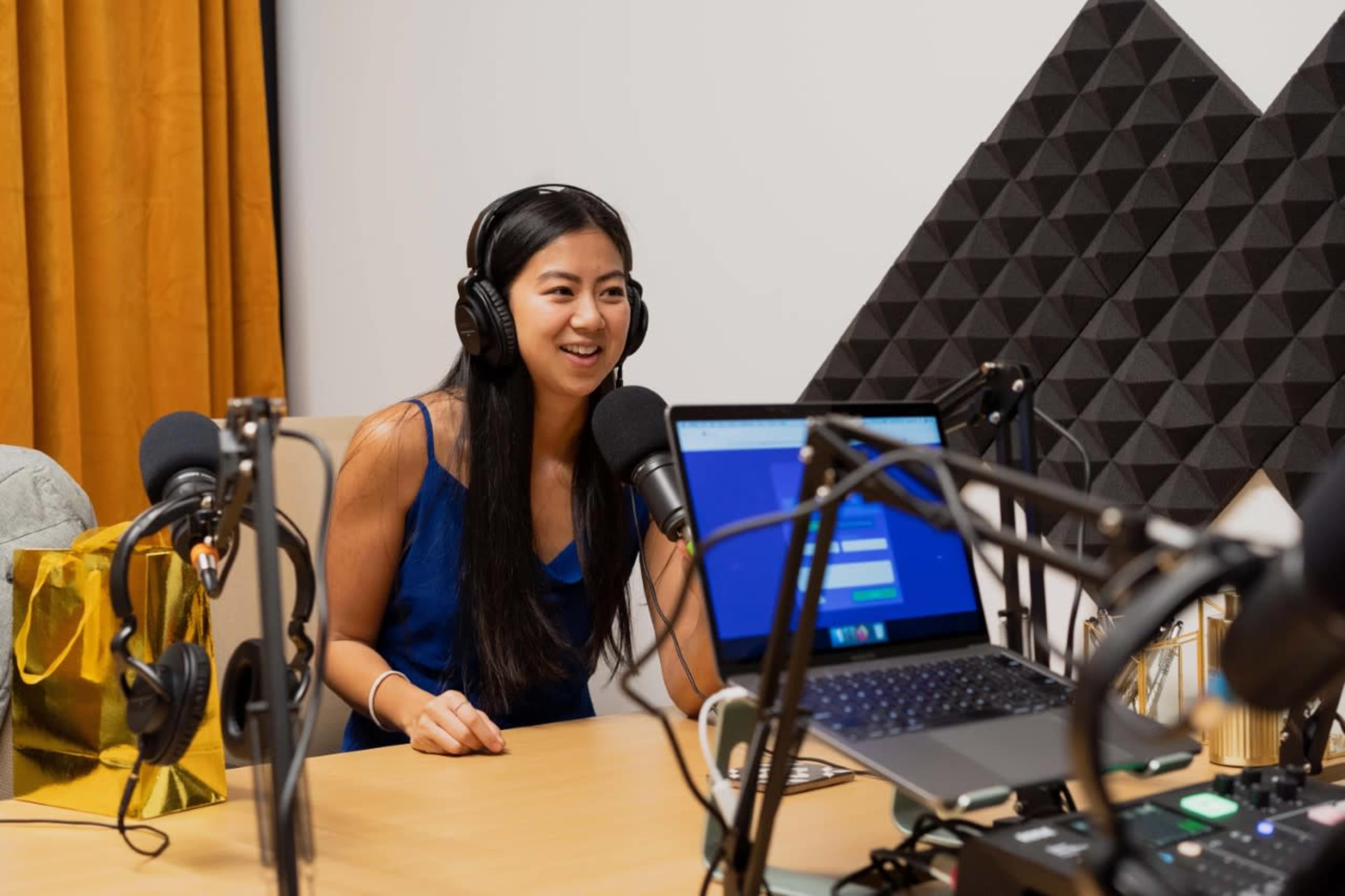 A woman wearing headphones sits at a table with microphones and a laptop in a podcast recording studio.