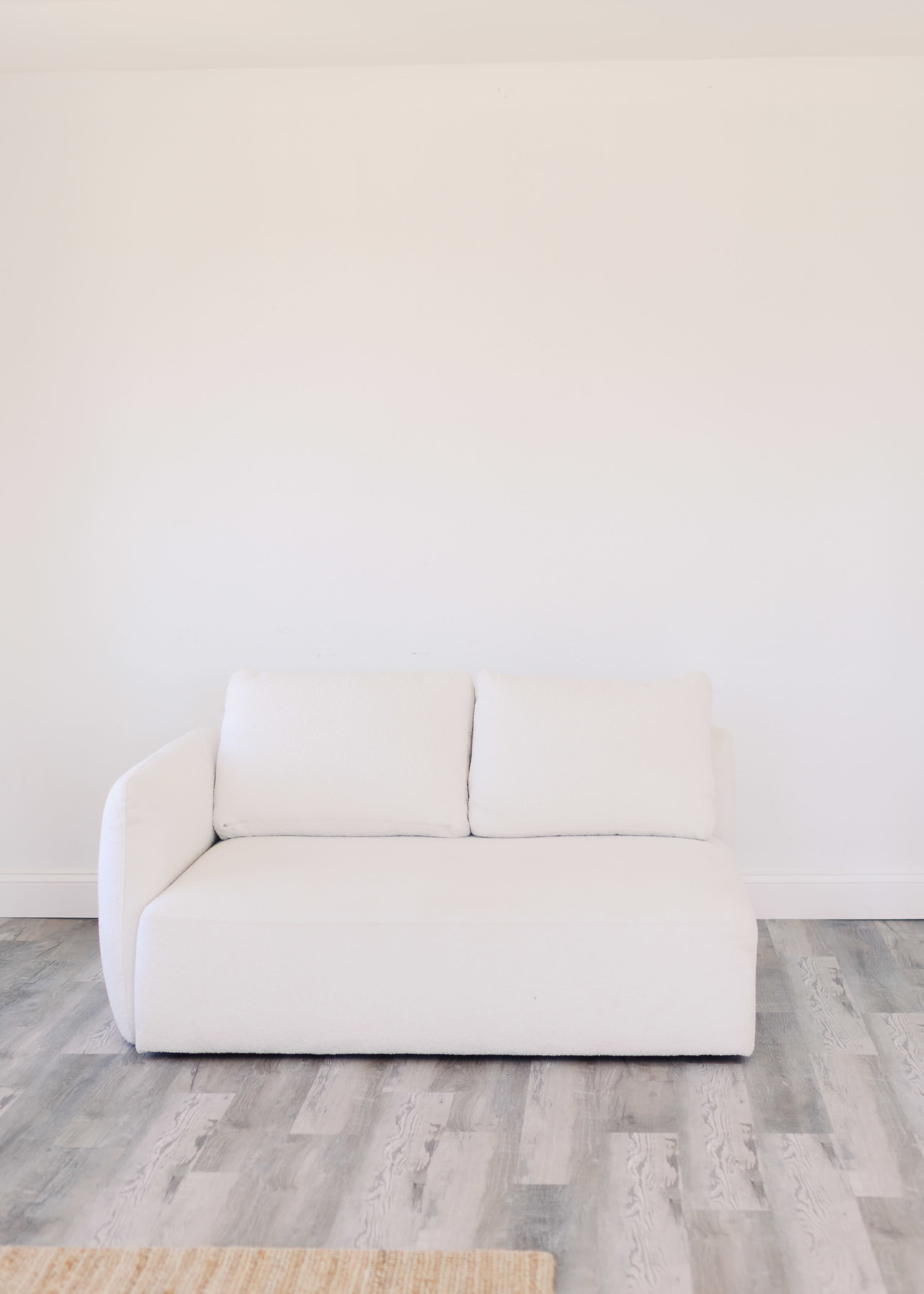 A white, minimalist two-seater couch is positioned against a plain white wall on wooden flooring.