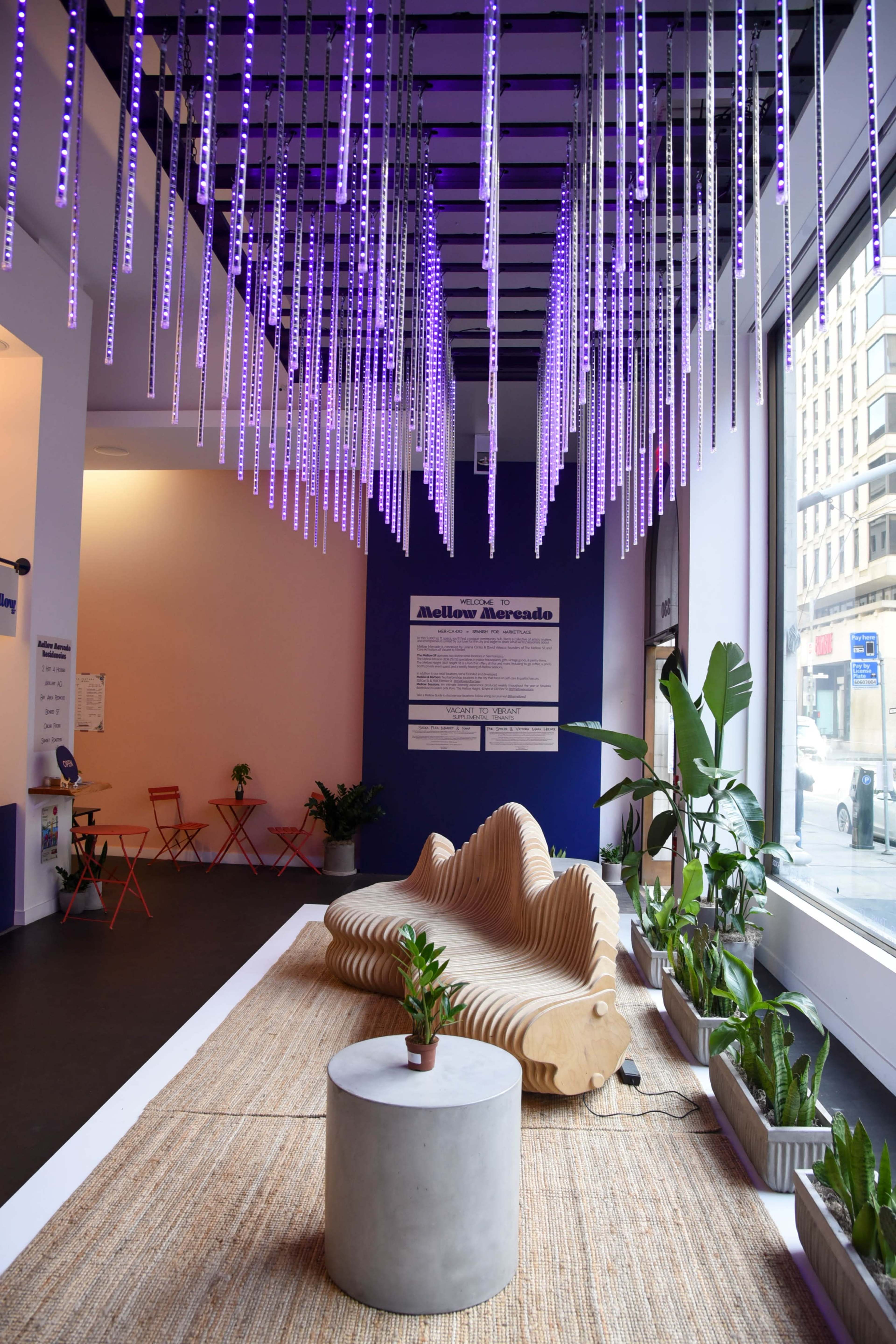 A modern interior space with a wavy wooden seating area, purple LED lighting hanging from the ceiling, and potted plants arranged along the floor.