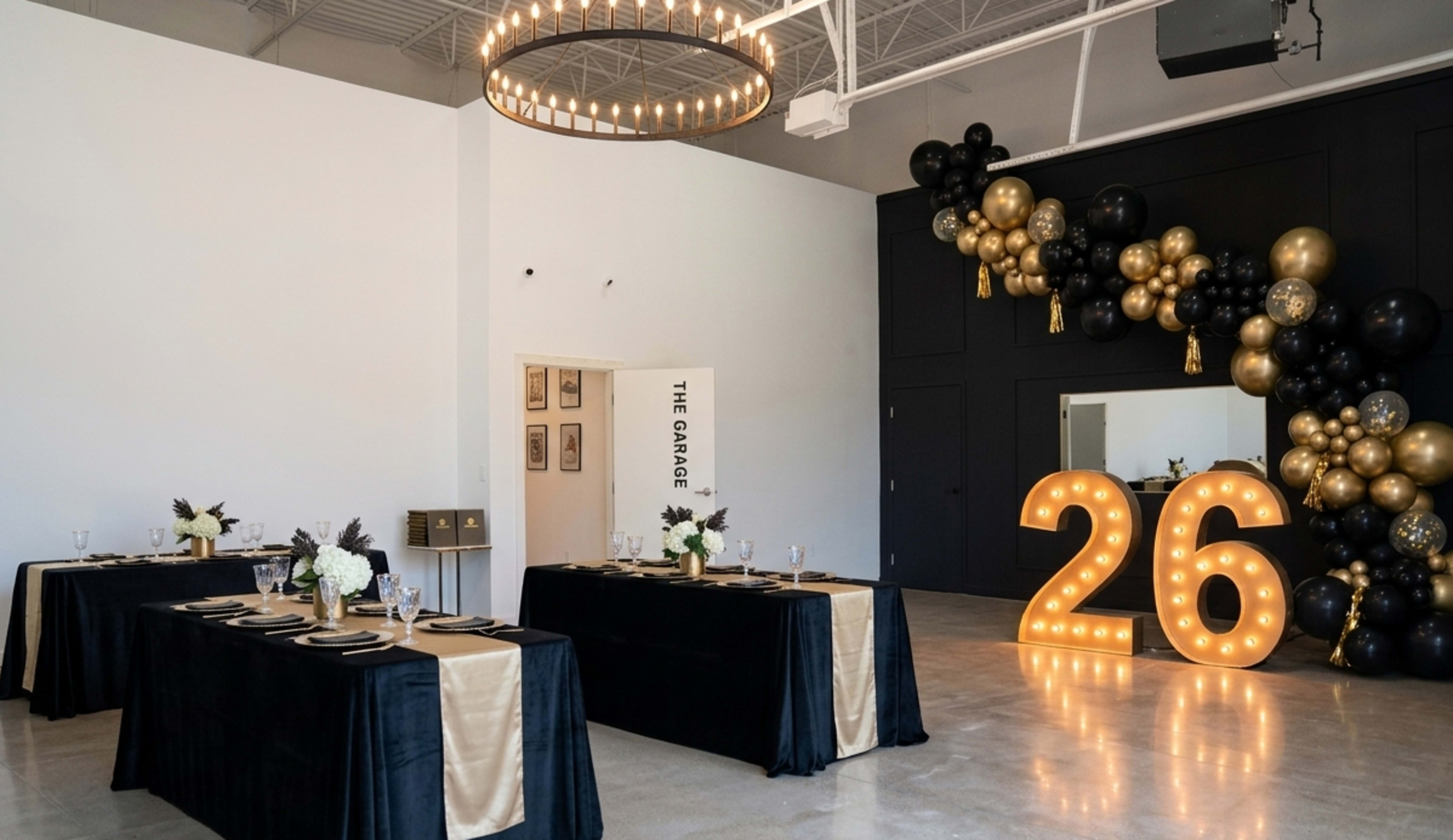 The image shows a spacious event venue with two long tables set with black tablecloths and gold accents, illuminated by a large chandelier and decorated with a balloon arrangement.