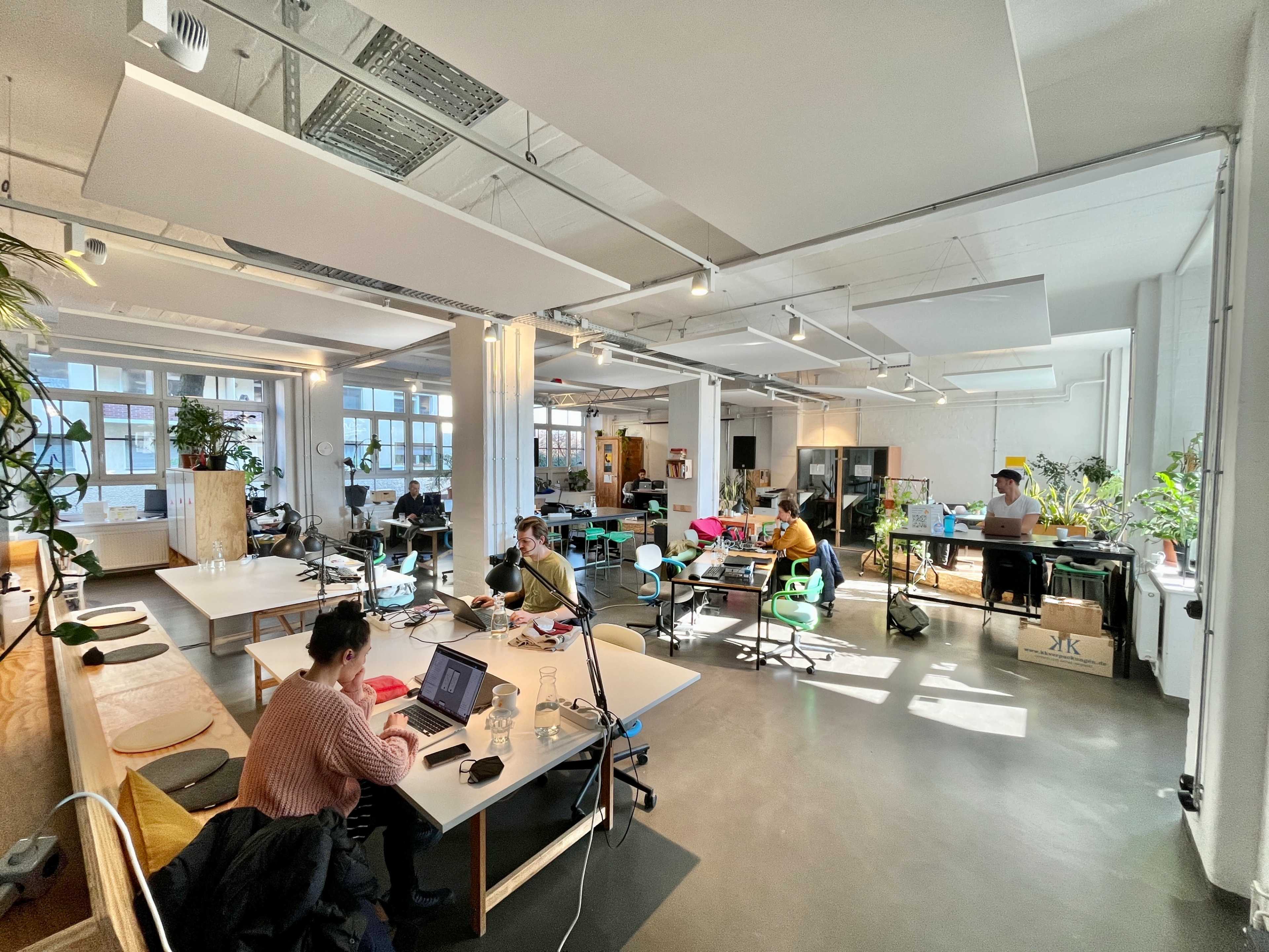 A spacious office environment with multiple workstations, natural light streaming in through large windows, and several people engaged in various tasks.