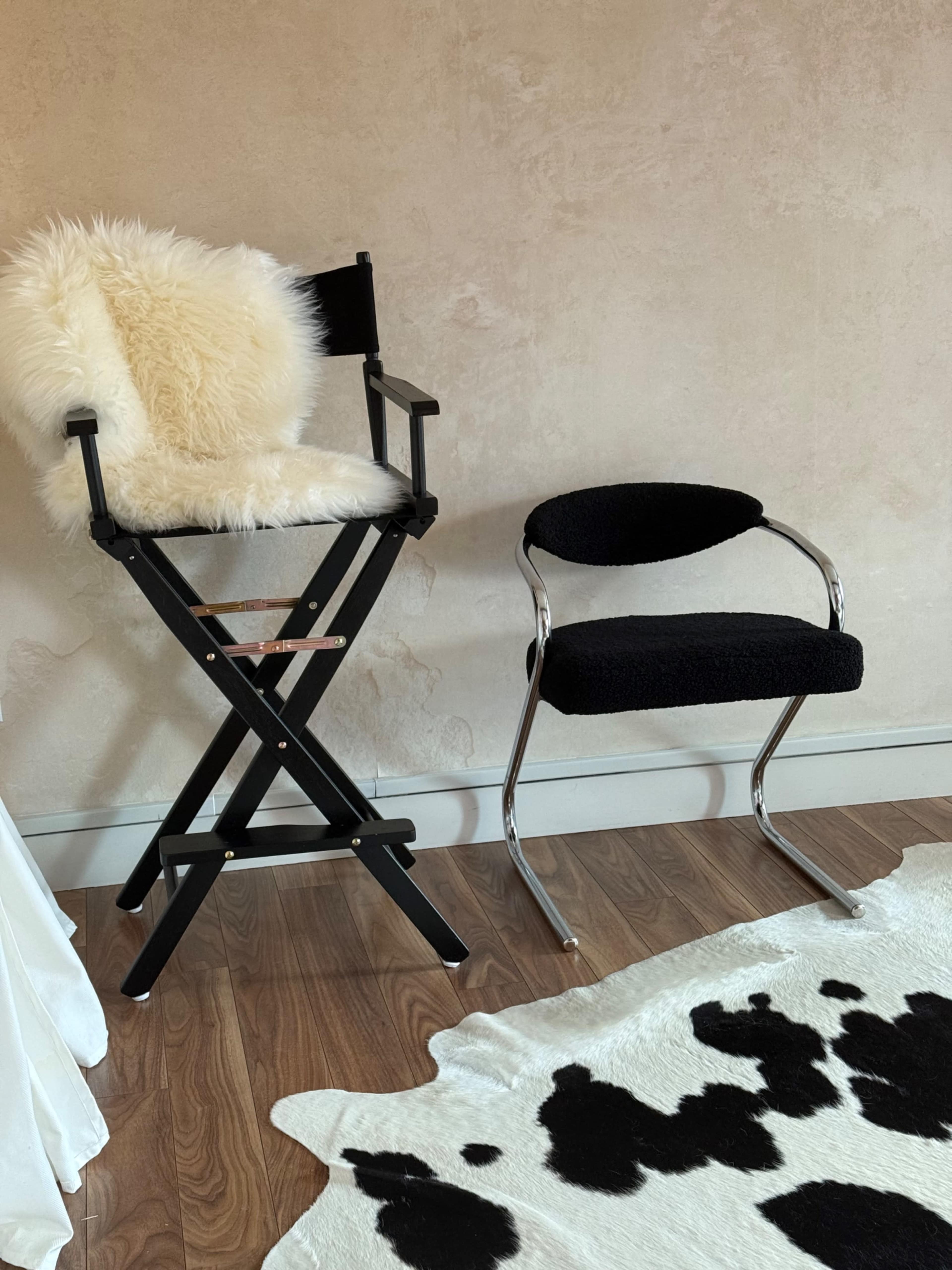 The image features a black director's chair with a cream-colored fur throw beside a minimalist black chair, both positioned next to a cow-print rug on wooden flooring.