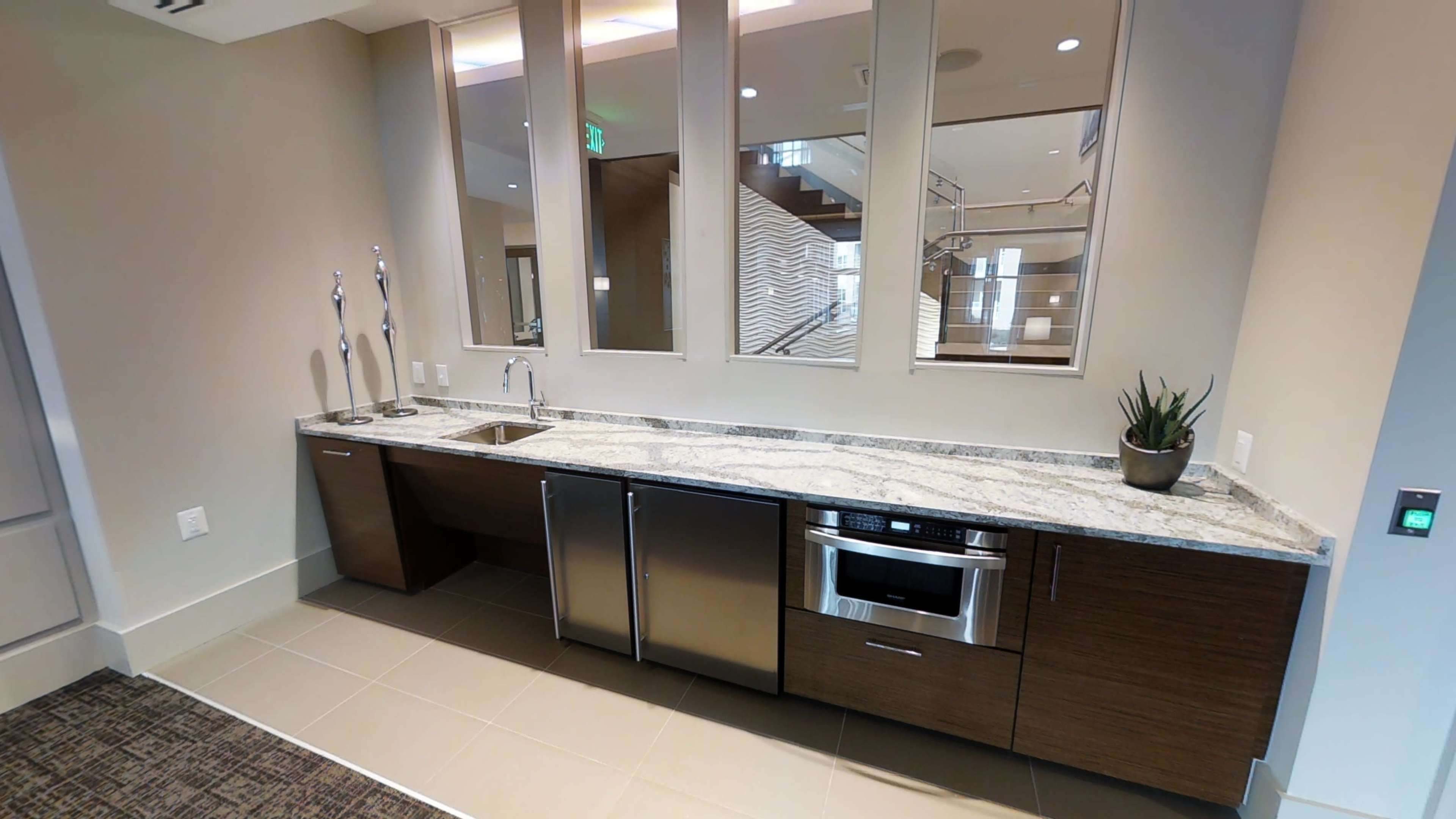 The image shows a modern kitchen area featuring a long countertop with a sink, stainless steel appliances, and a decorative plant, all set against a neutral wall with large windows.
