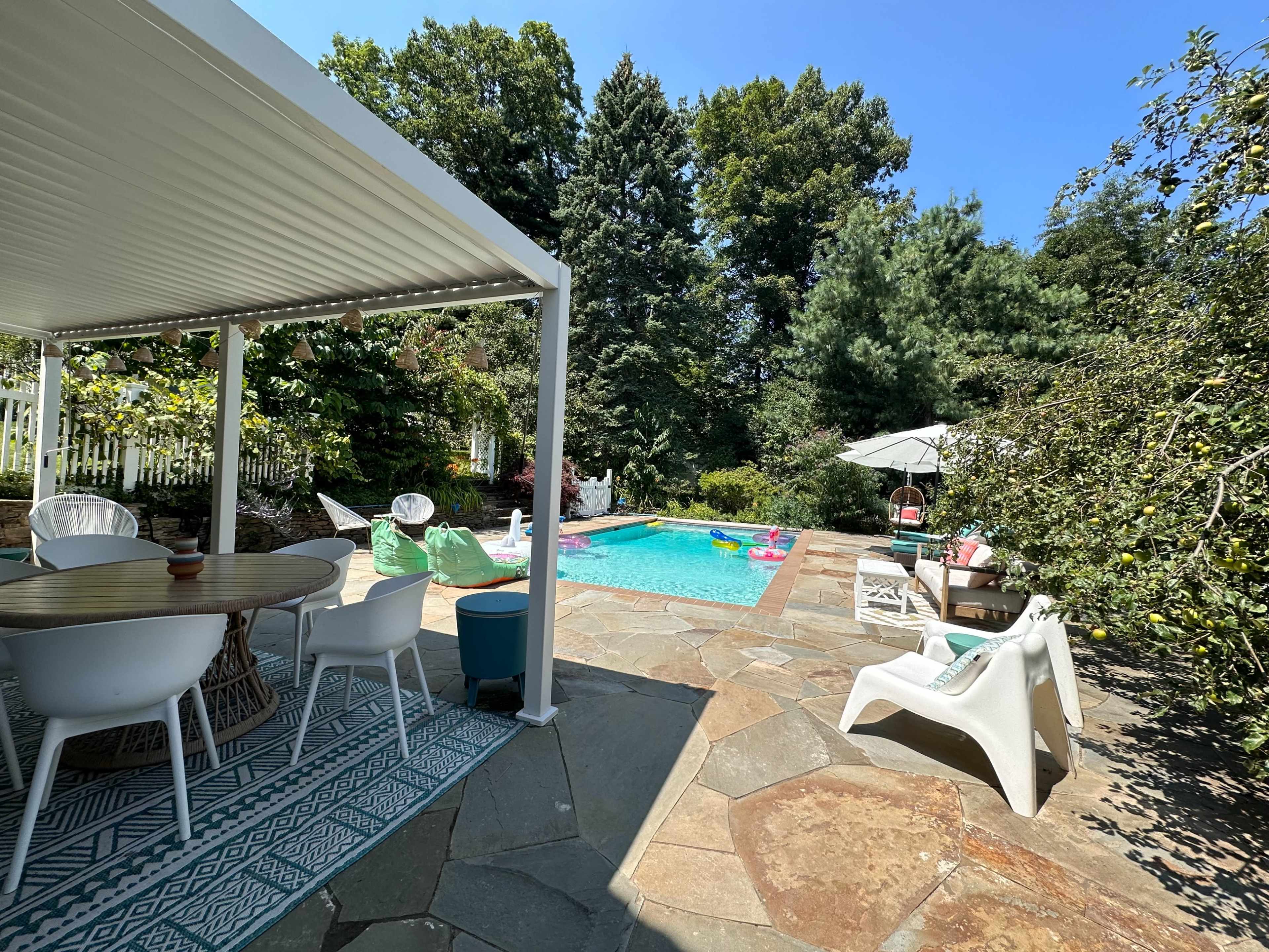 A patio area featuring a dining table and chairs overlooks a swimming pool surrounded by greenery and lounge chairs.