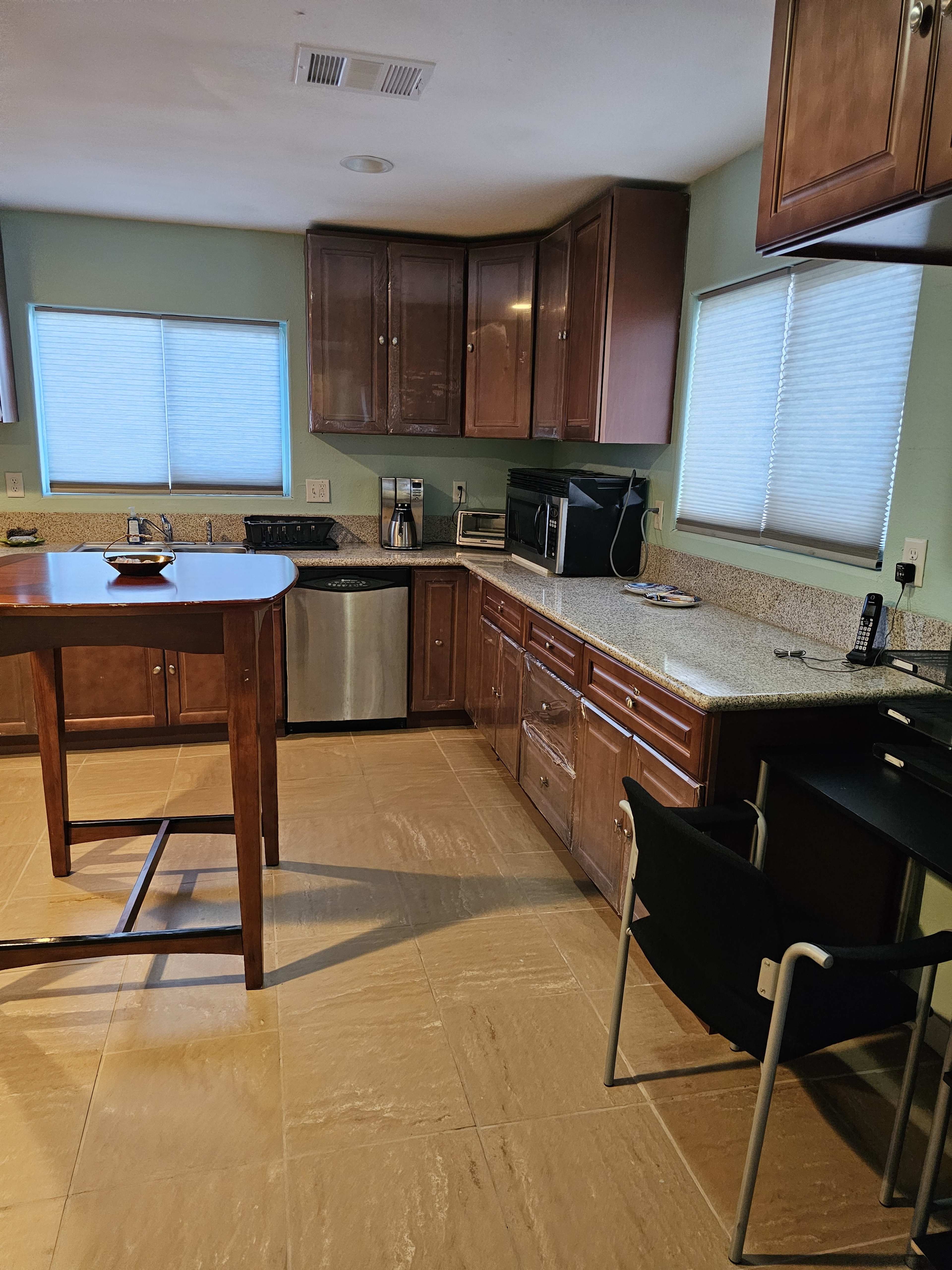 A kitchen features wooden cabinets, granite countertops, a stainless steel sink, and appliances including a microwave and coffee maker, with a table and a chair positioned nearby.