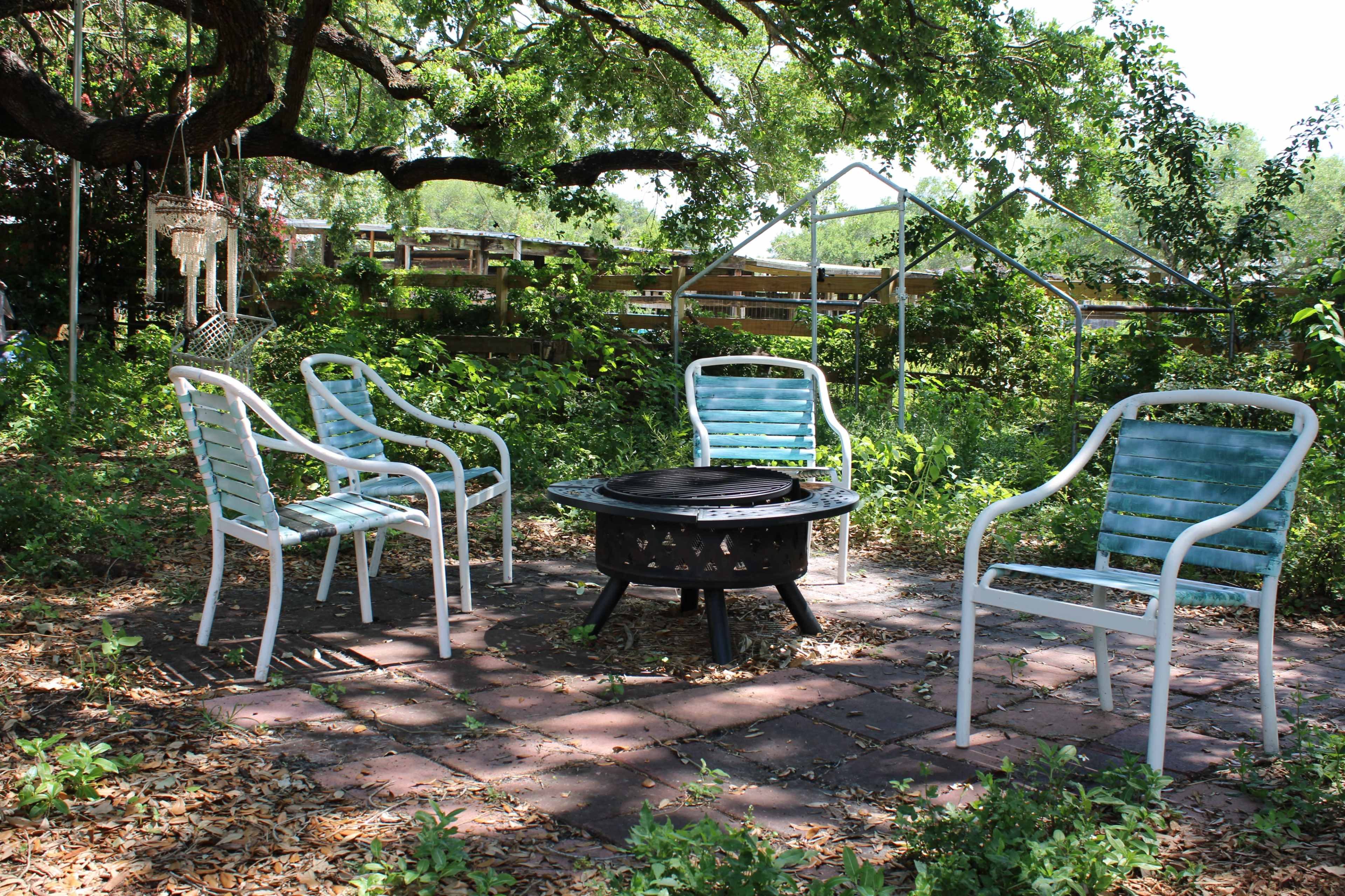 A fire pit surrounded by four white metal chairs is set in a grassy area with lush greenery and scattered fallen leaves.