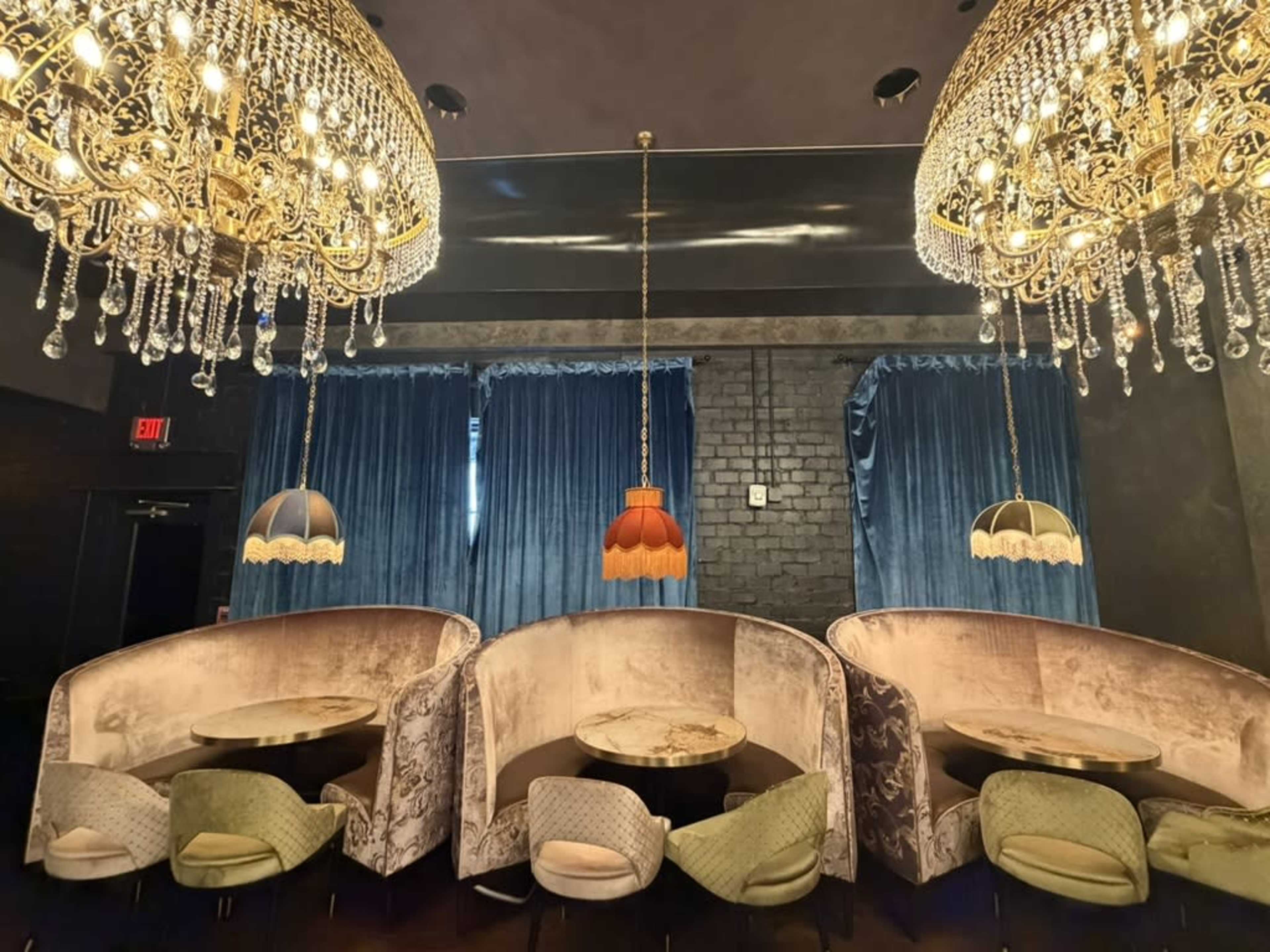 The image shows a stylish interior of a restaurant or lounge featuring plush seating, ornate chandeliers, and rich blue drapery.