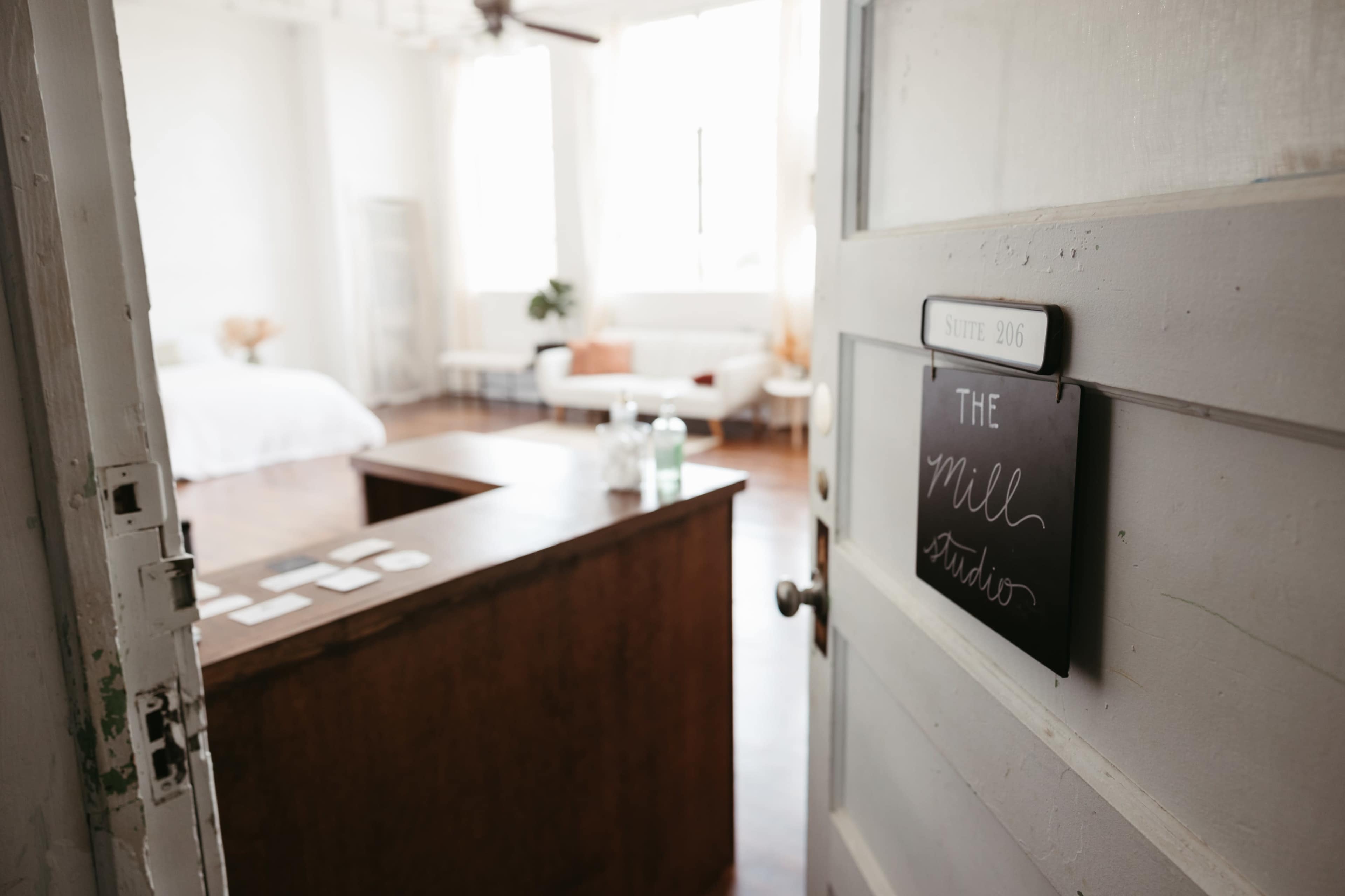 A wooden door with a sign reading "The Mill Studio" leads into a spacious, well-lit room featuring a bed, seating area, and plants.