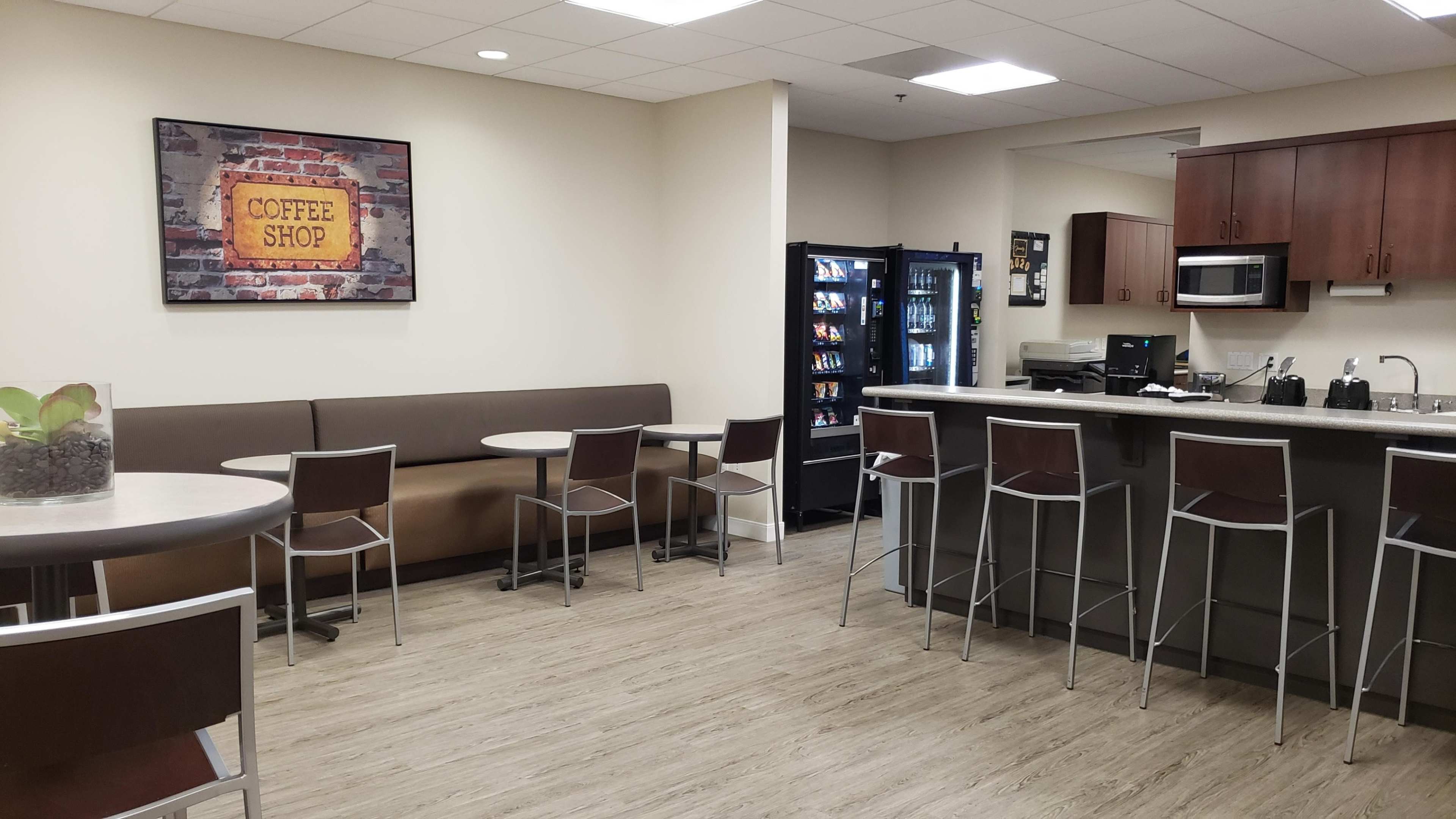 The image shows a small café area with seating, a drink vending machine, and a counter space in a well-lit room.