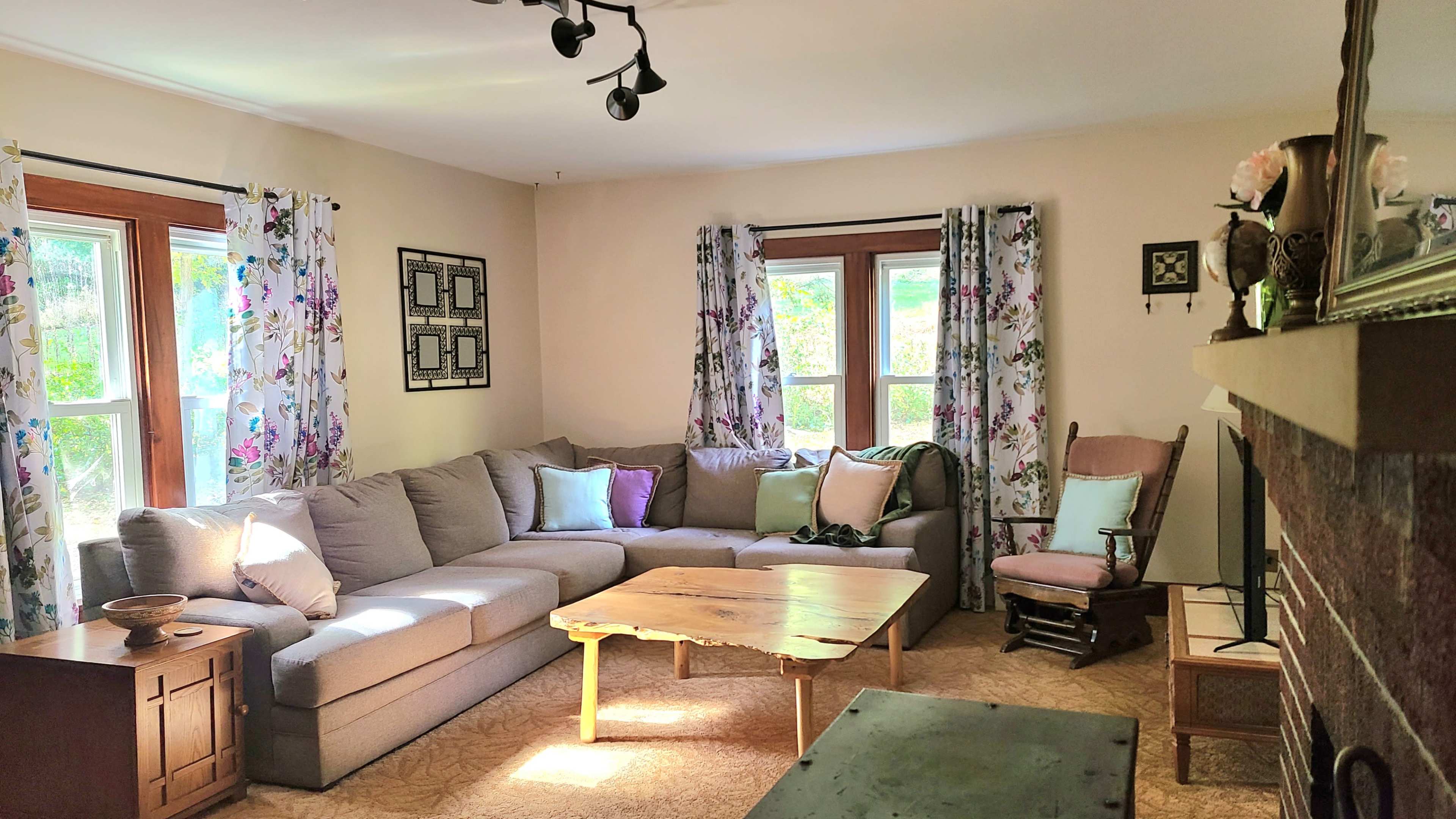 The living room features a large sectional couch, a wooden coffee table, a rocking chair, and floral curtains on the windows.