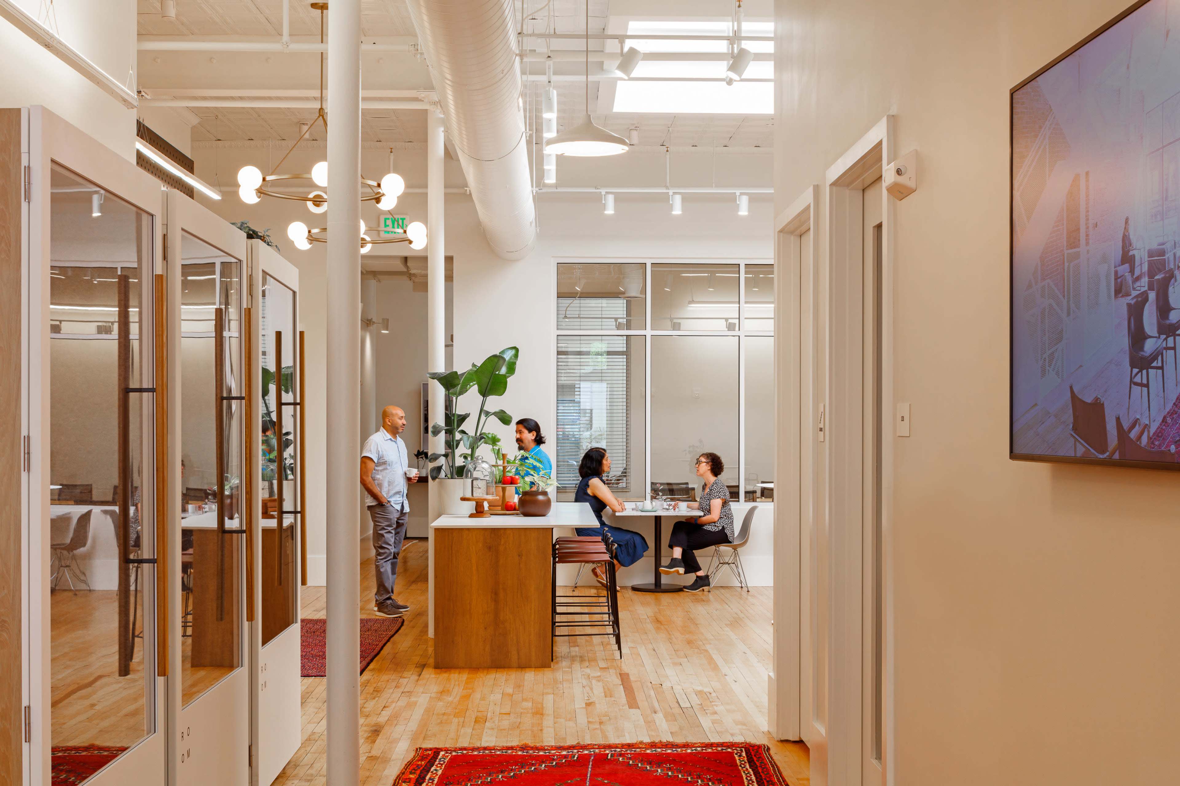 A well-lit modern office features two distinct areas: a small informal meeting space with two people at a table and a second area where two individuals converse near a large countertop.