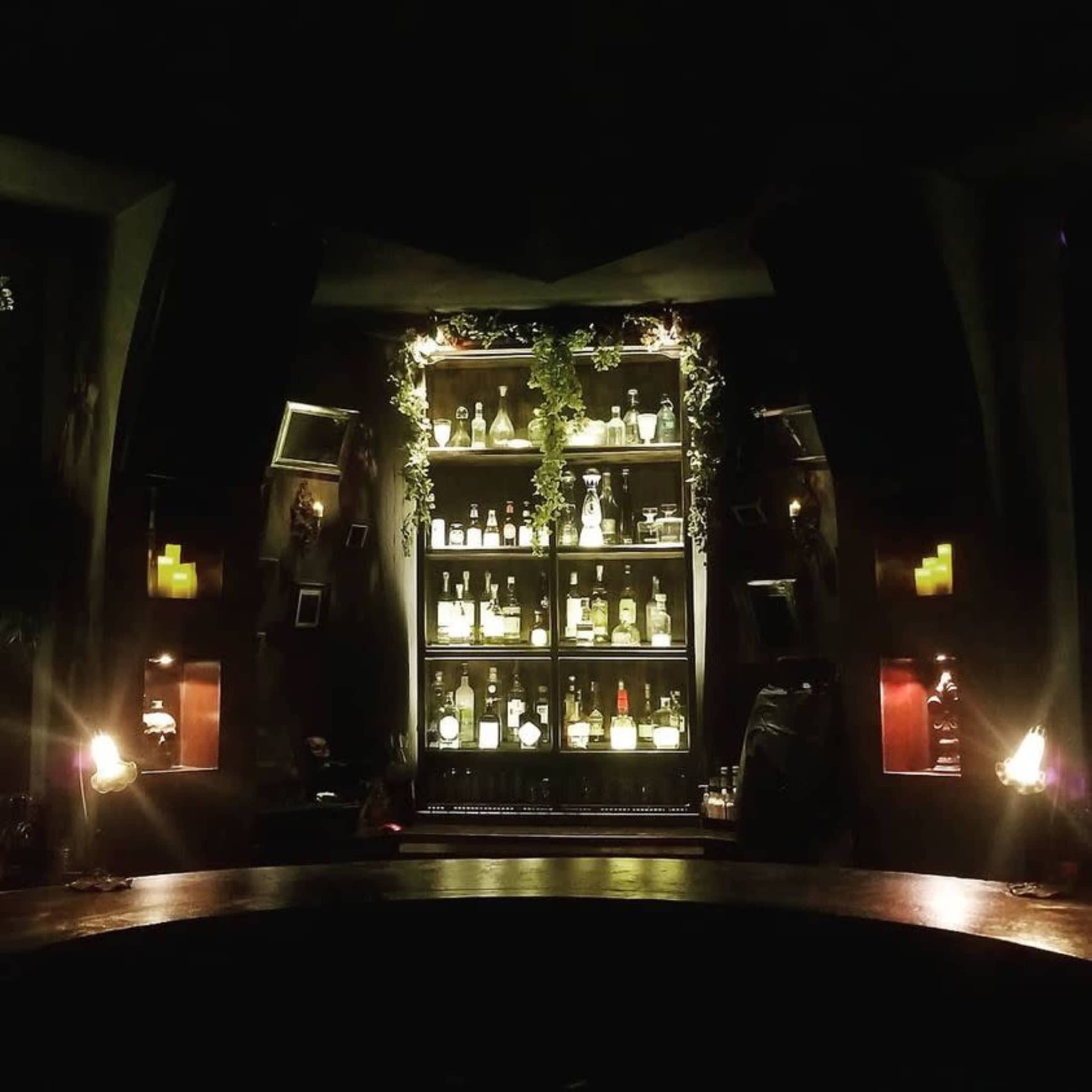 The image shows a dimly lit bar with a shelf filled with various bottles and decorative elements, set against a dark backdrop.
