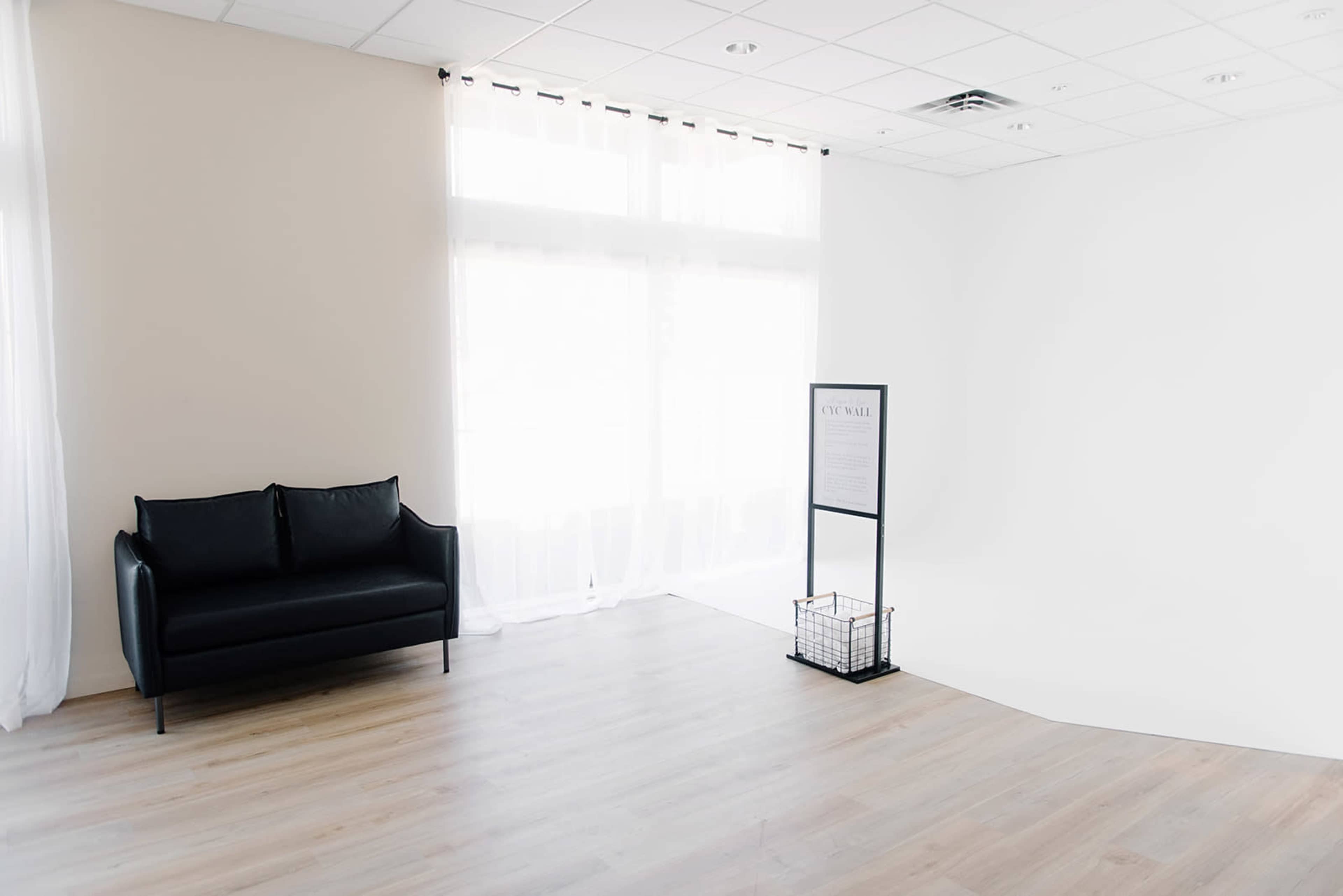 A minimalistic interior space features a black couch, a tall display stand, and large windows with sheer curtains, all set against light-colored walls and wooden flooring.