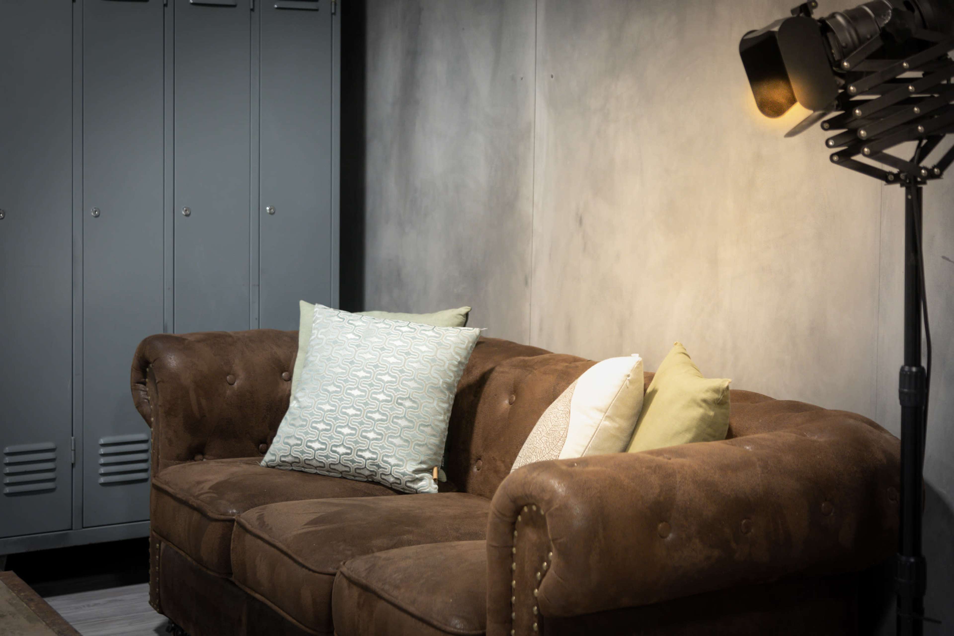 A brown leather couch with decorative pillows against a textured gray wall, accompanied by industrial-style lighting and lockers in the background.