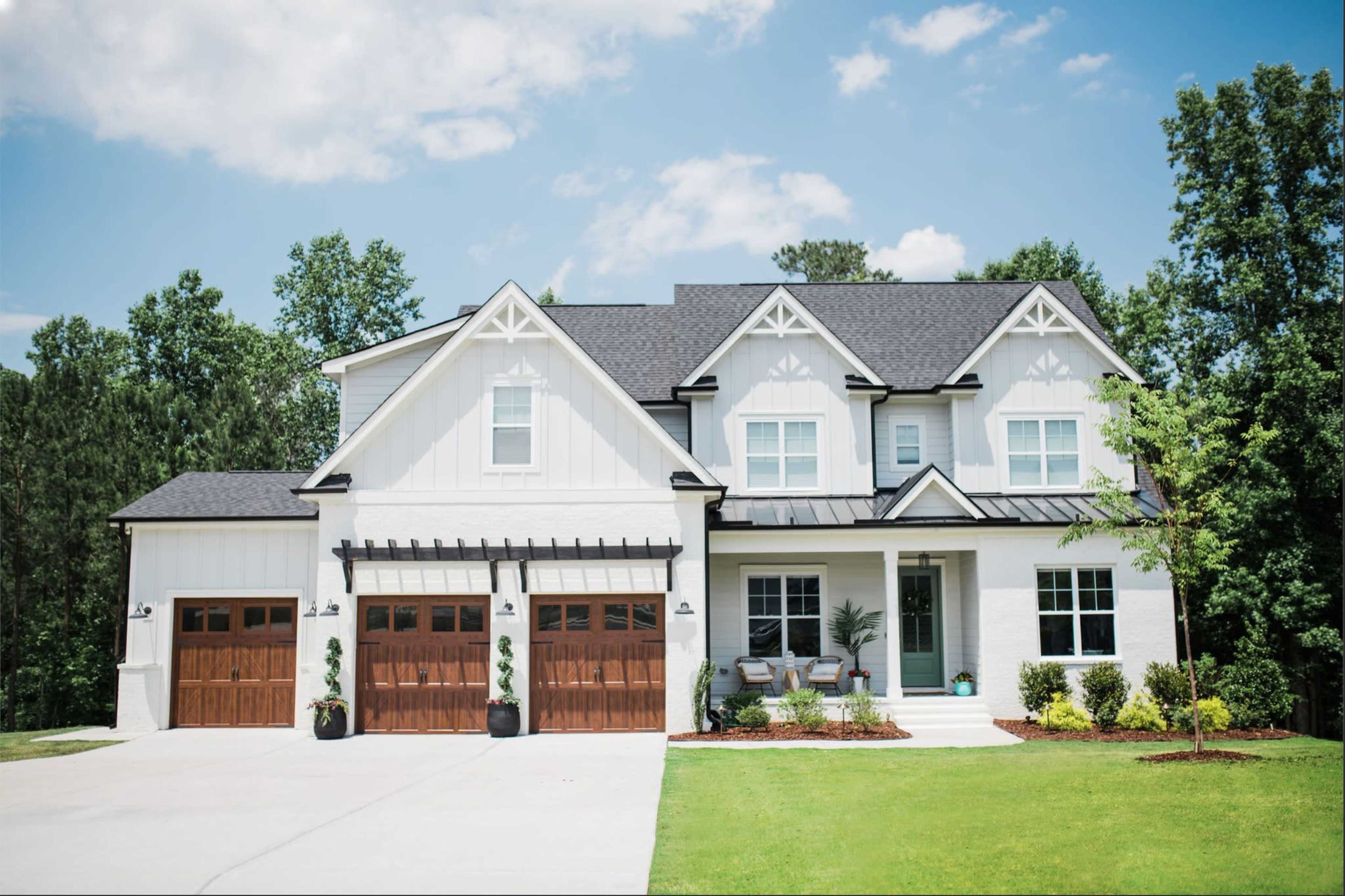 A two-story white house with a black roof and wooden garage doors, surrounded by a well-manicured lawn and trees.