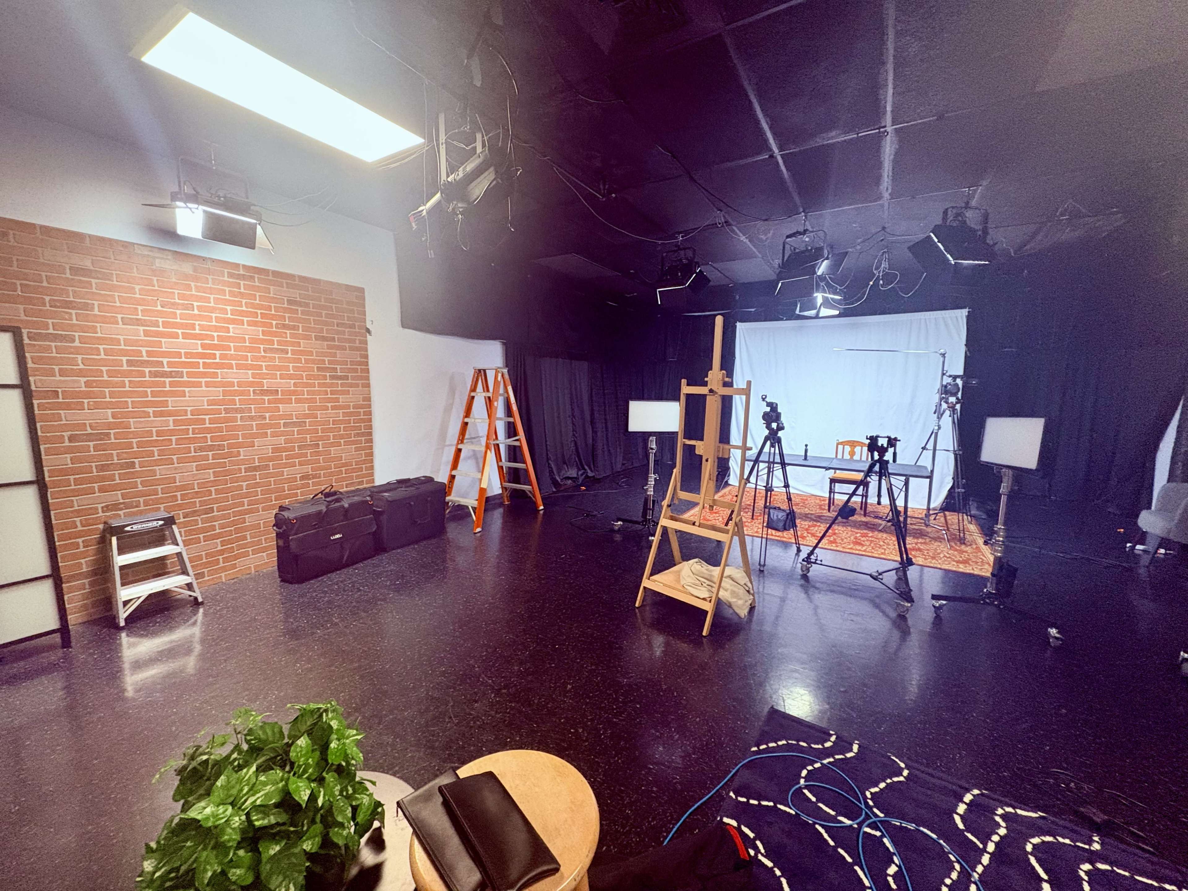 The image shows a spacious studio with a brick wall, lighting equipment, cameras on tripods, an easel, and various props arranged for a photoshoot or video production.