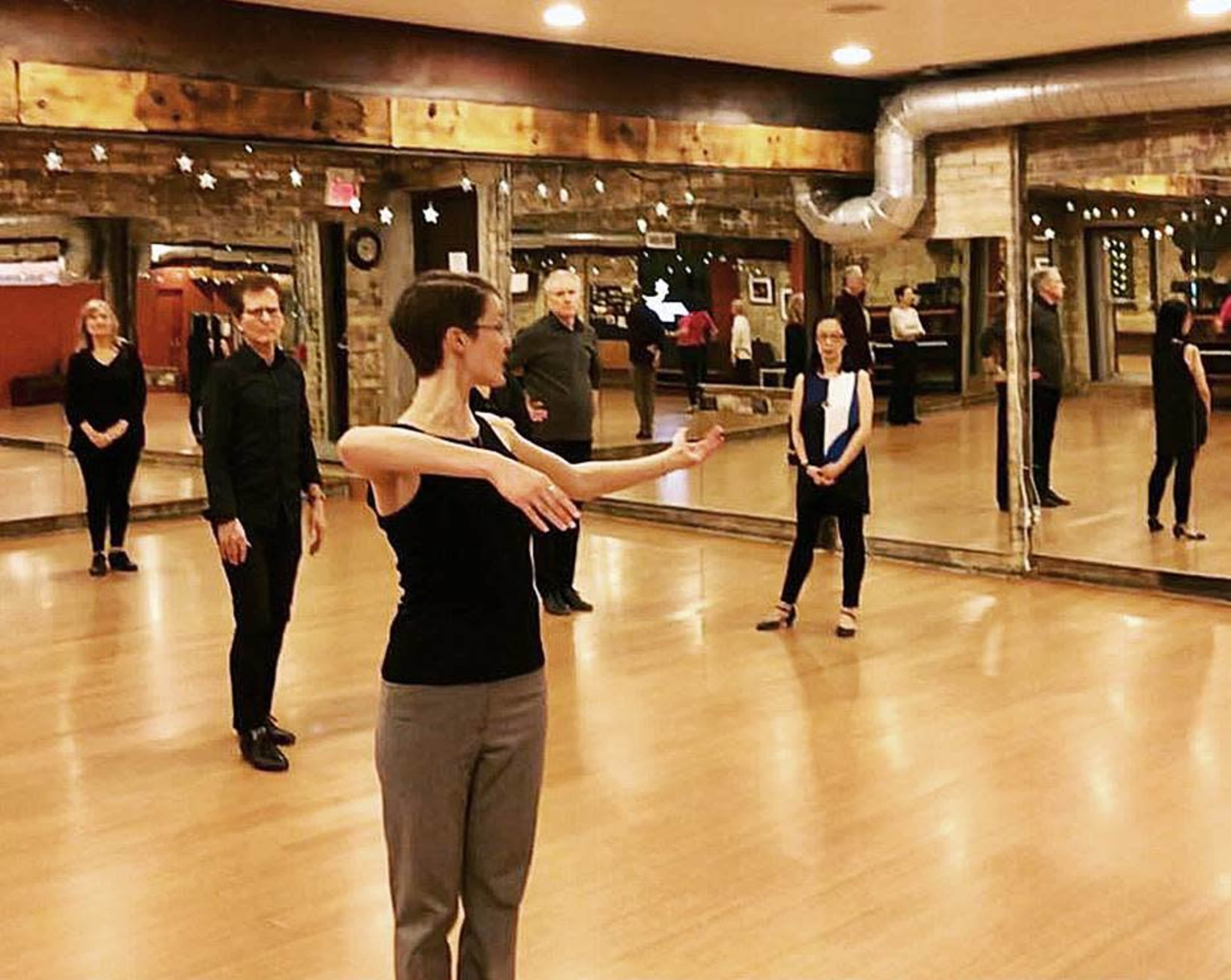 A dance instructor demonstrates a movement in a mirrored studio filled with students.