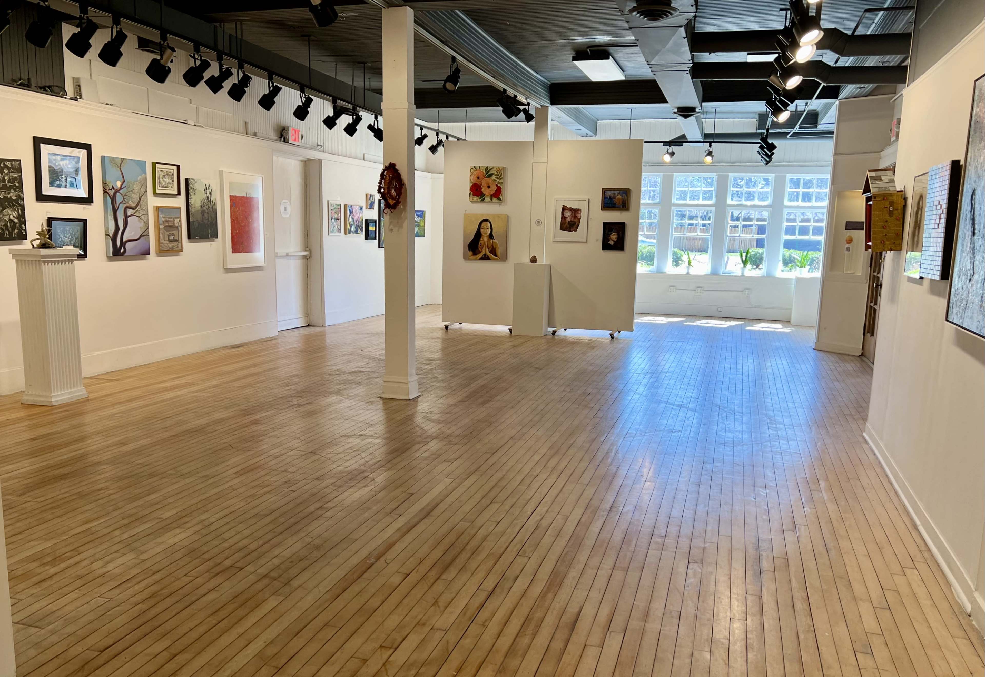 A spacious art gallery with wooden floors and white walls displaying various framed artworks, illuminated by overhead lighting.