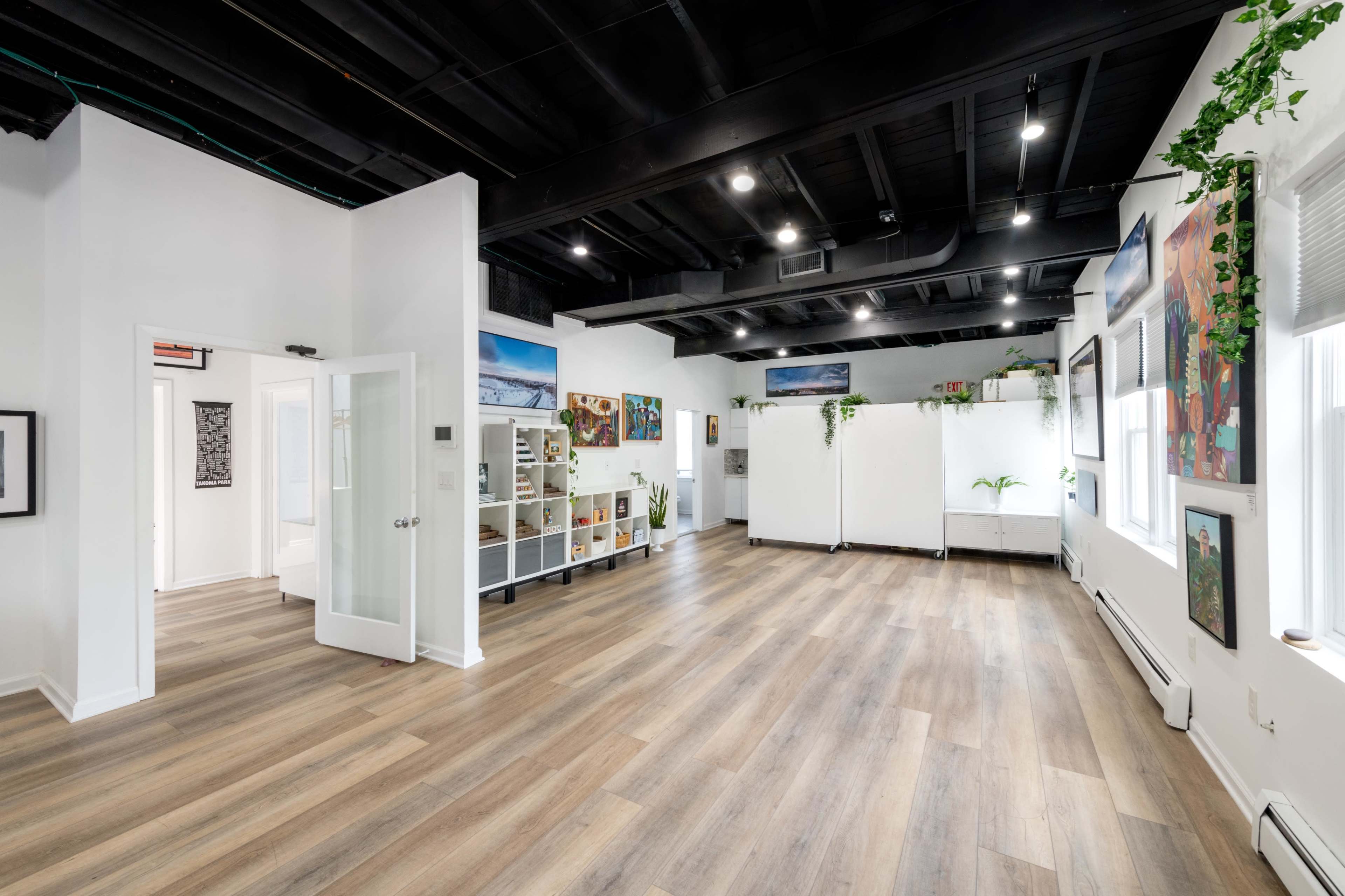 A spacious, well-lit room with wooden flooring, white walls, and plants, featuring artwork on the walls and a shelf along one side.
