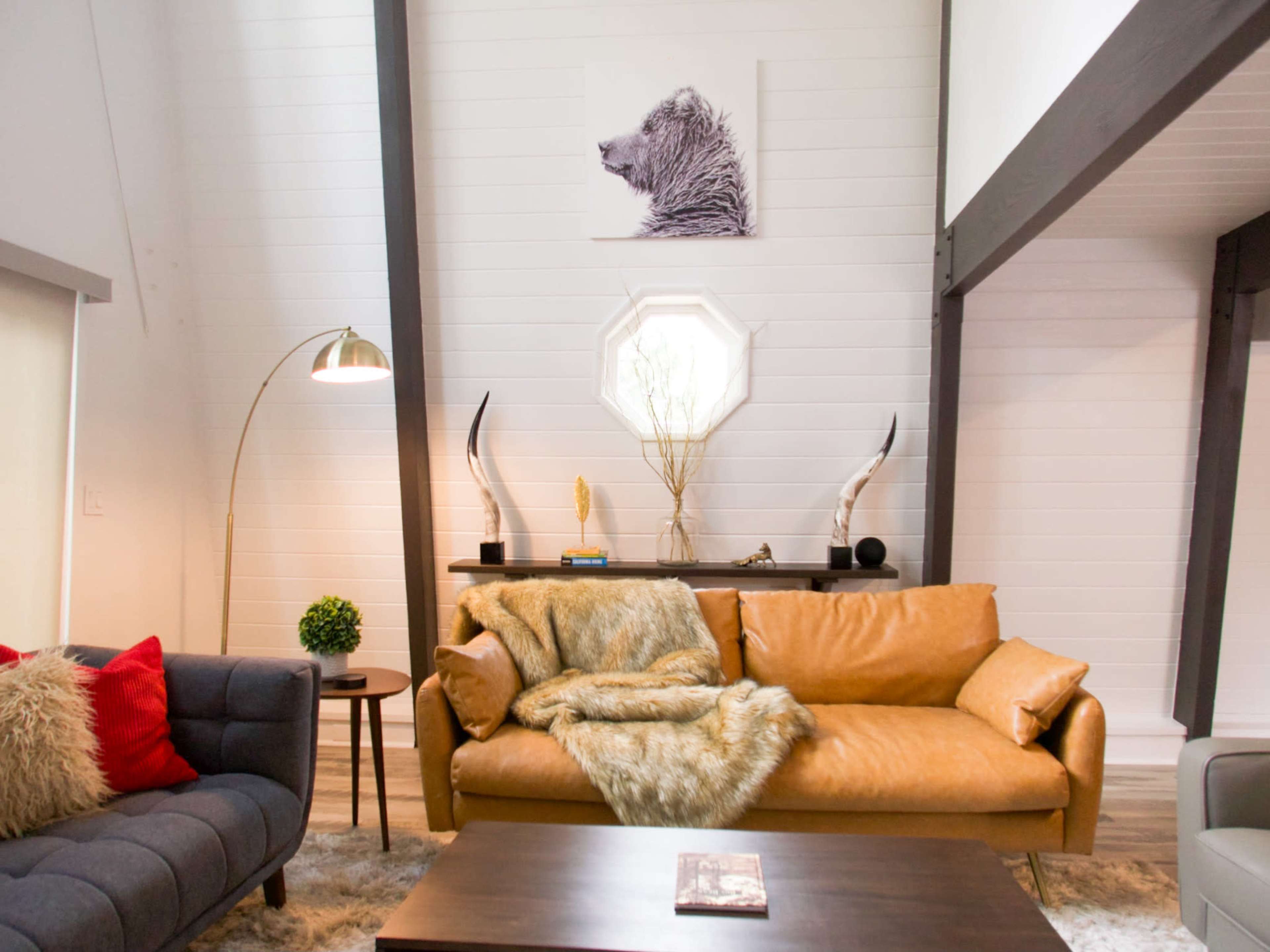 The living room features a brown leather sofa, a gray upholstered couch, and a decorative bear portrait on the wall above a console table with antlers and a plant.