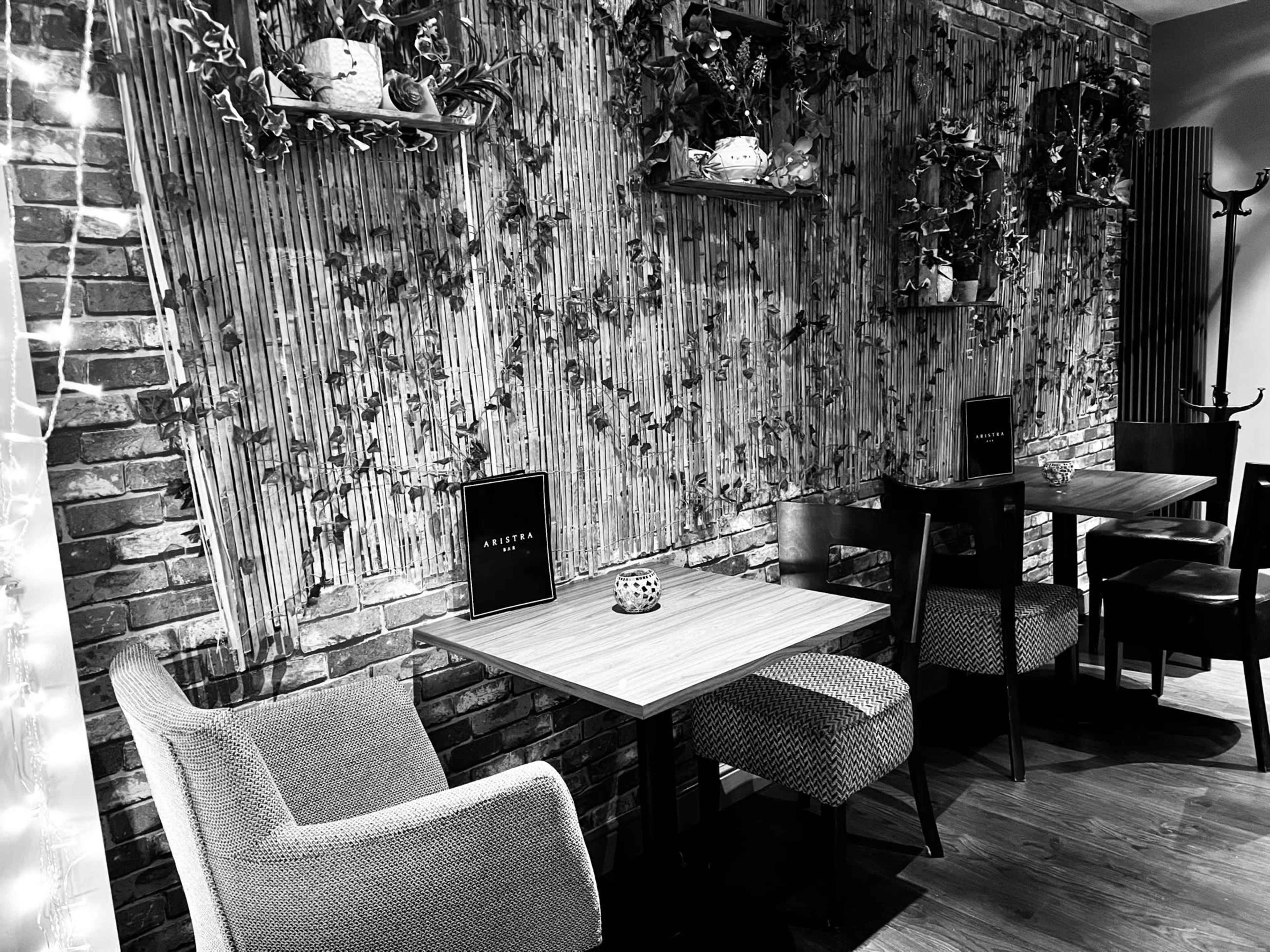 The image shows a cozy corner of a café with wooden tables, chairs, and a decorative wall featuring plants and bookshelves.