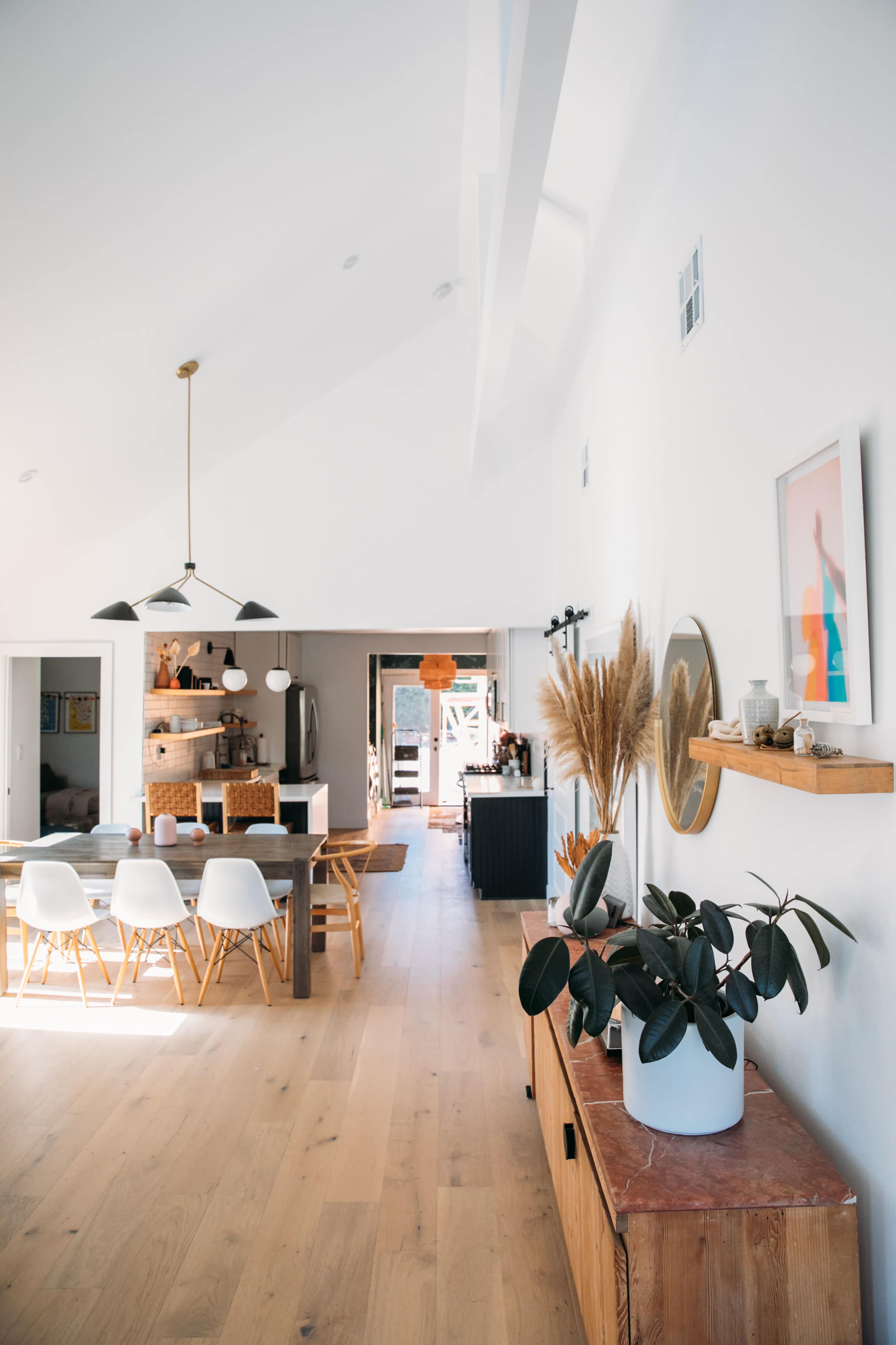 A bright, modern open-concept living space features a dining area with a large wooden table and white chairs, leading to a kitchen and outdoor access.