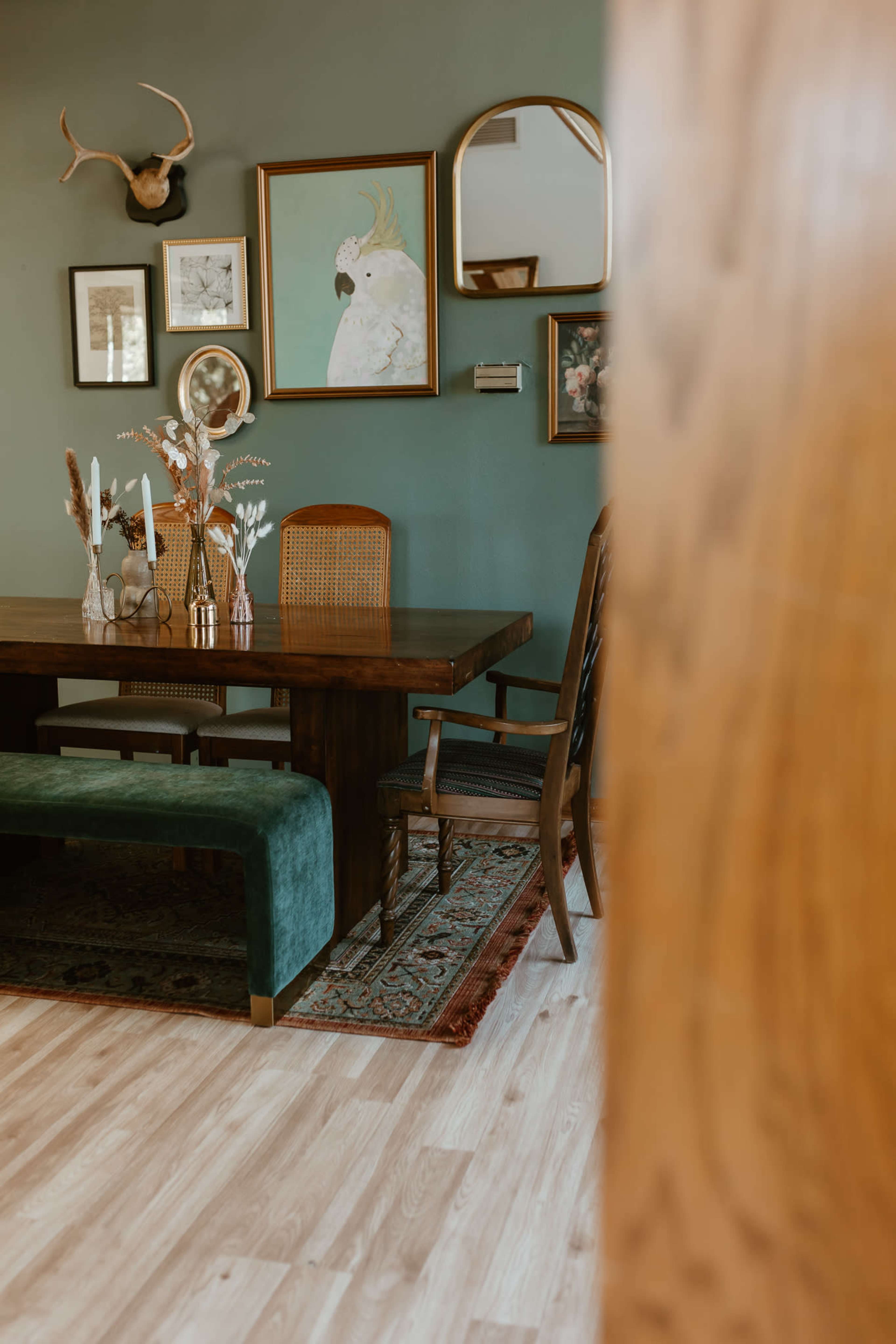 A dining area features a wooden table surrounded by various chairs, decorated with framed artwork on green walls and a patterned rug beneath.