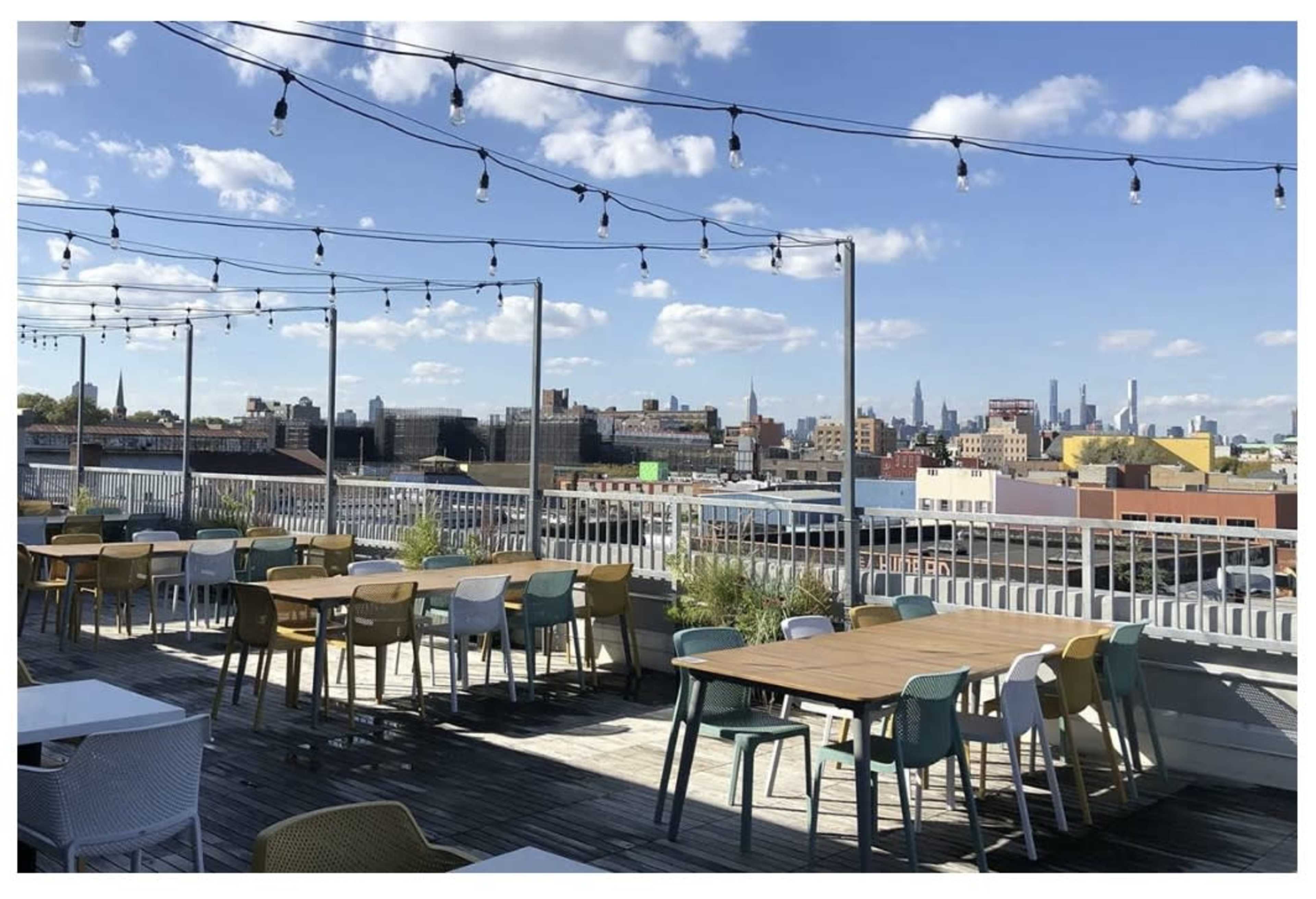 The image shows a rooftop terrace with several tables and chairs, string lights overhead, and a skyline view of buildings under a blue sky.