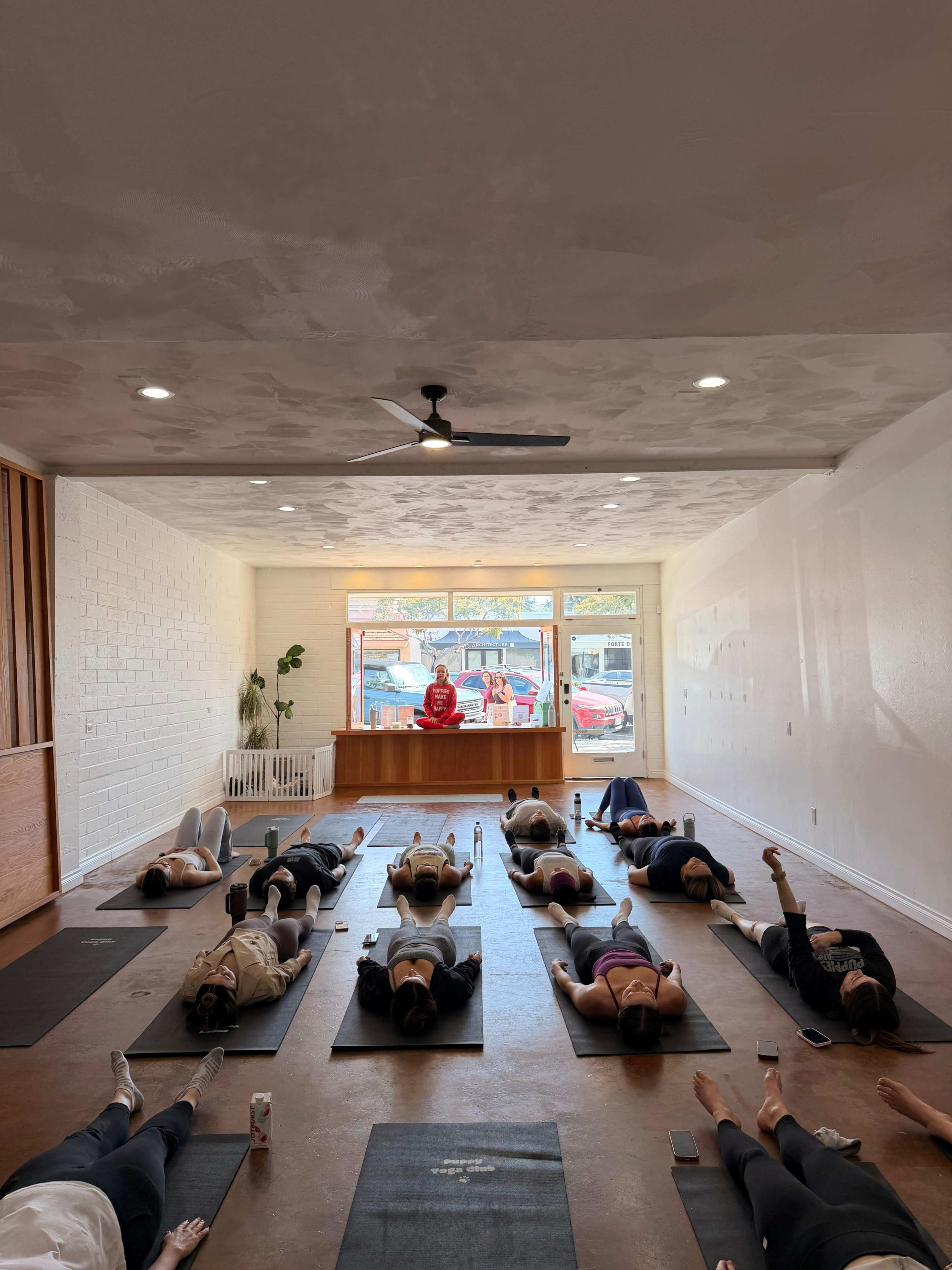 The image shows a group of people lying on yoga mats in a studio with a person standing at the front near a counter.