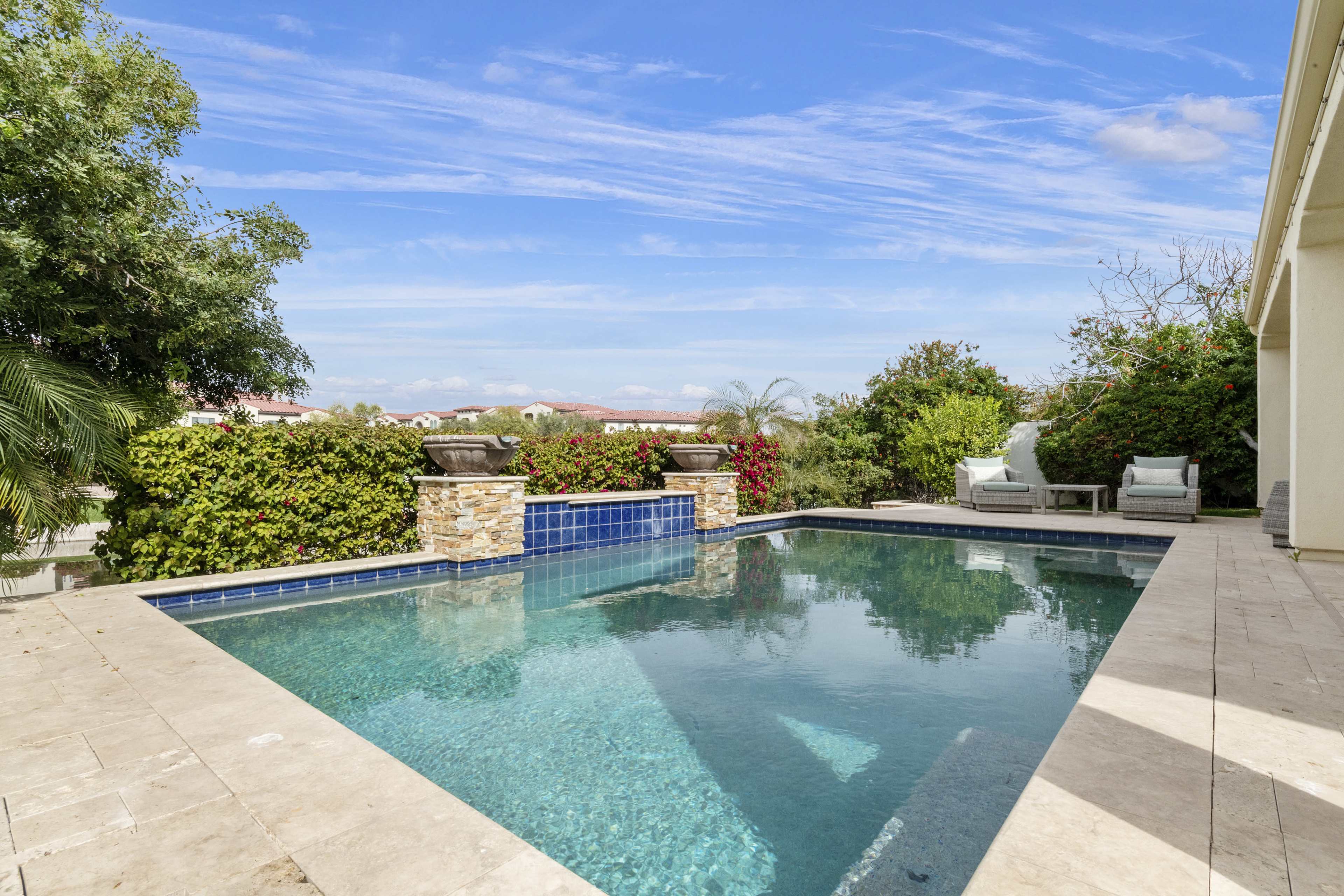 The image shows a clear swimming pool surrounded by a stone deck and lush greenery under a blue sky with scattered clouds.