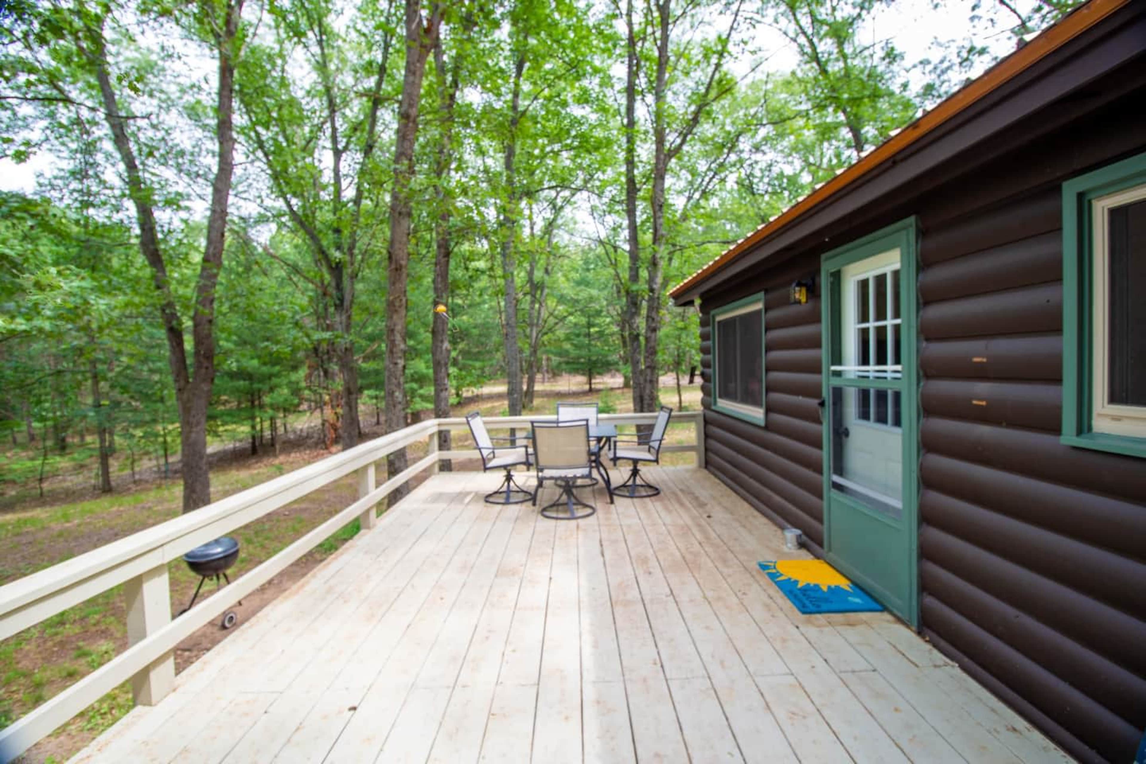 A wooden deck with a table and chairs overlooks a wooded area beside a cabin.