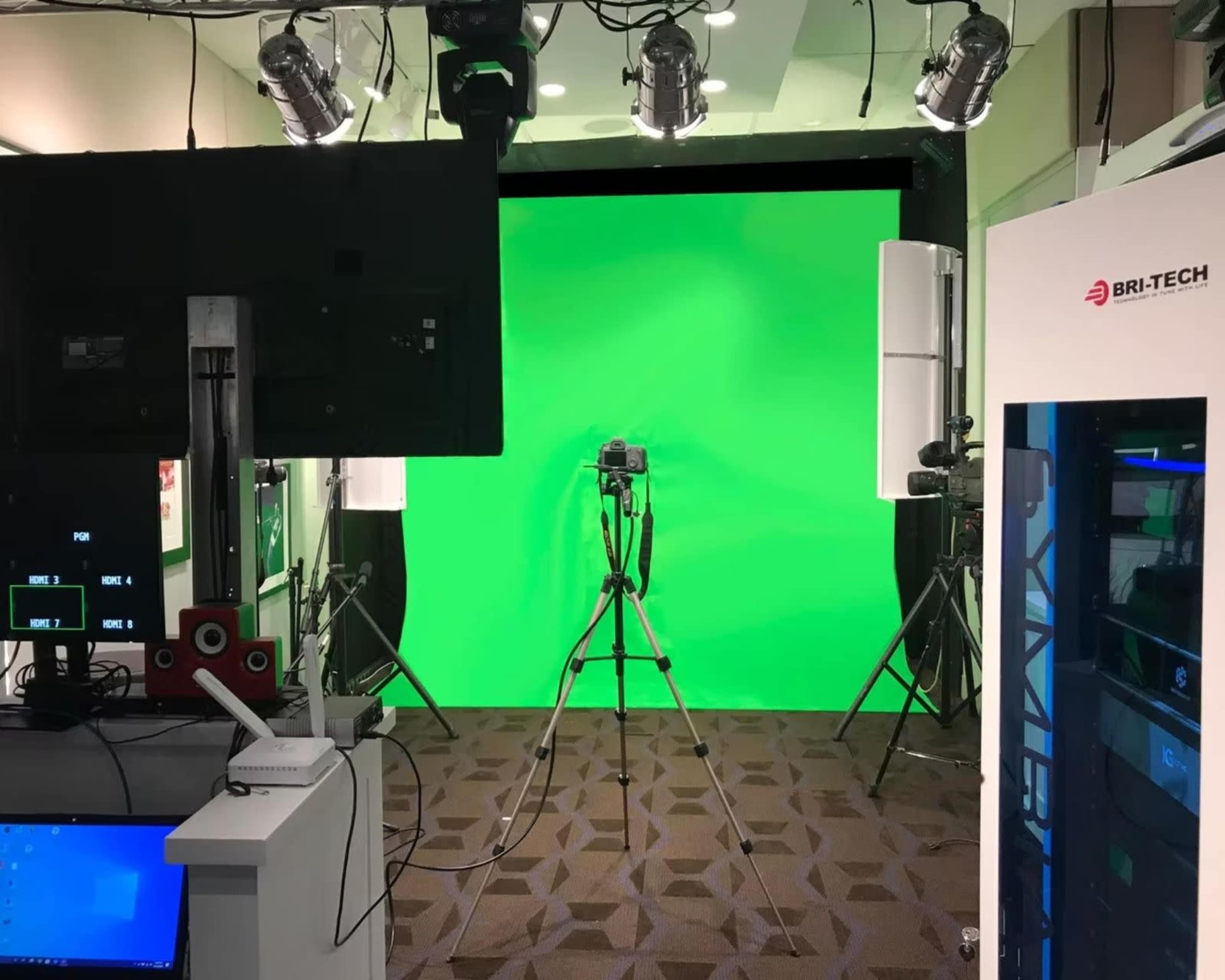 The image shows a green screen setup in a studio, equipped with lights and a camera on a tripod, alongside a computer and technical equipment.