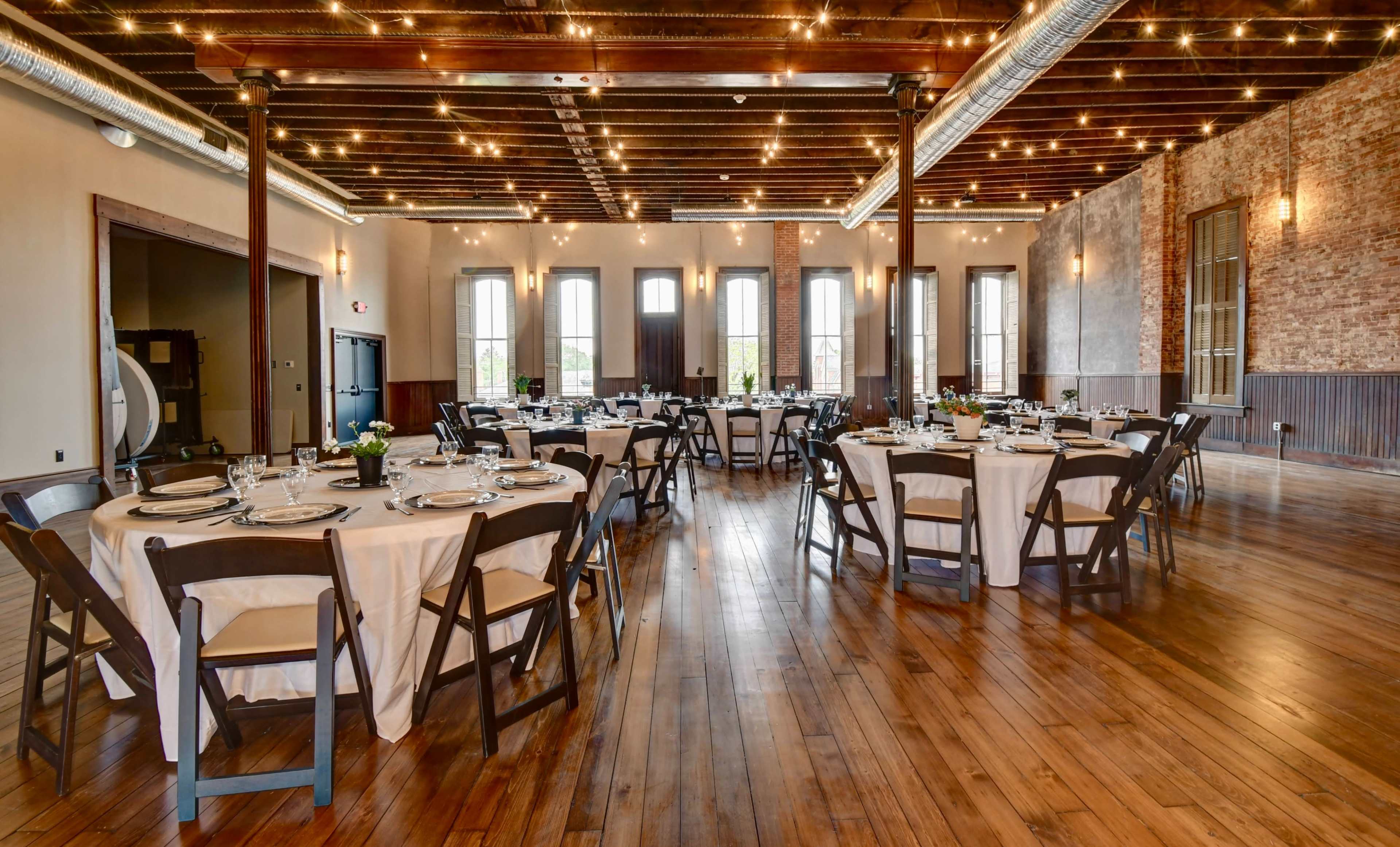 A spacious dining area with round tables set for a meal, under string lights and featuring exposed wood beams and a brick wall.