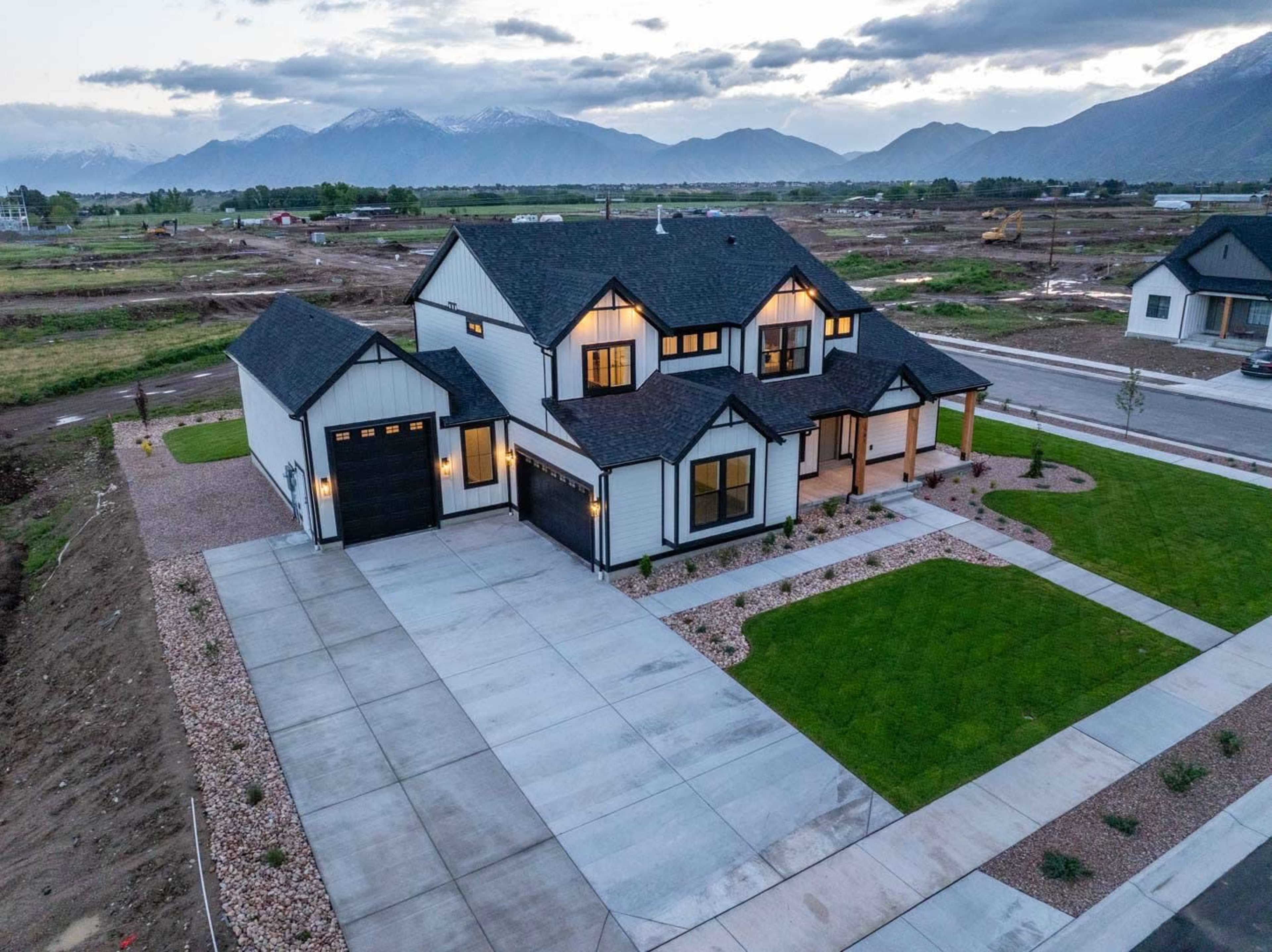A two-story modern house with a dark roof and light-colored exterior is situated on a well-manicured lawn, surrounded by mountains and a partially developed landscape.