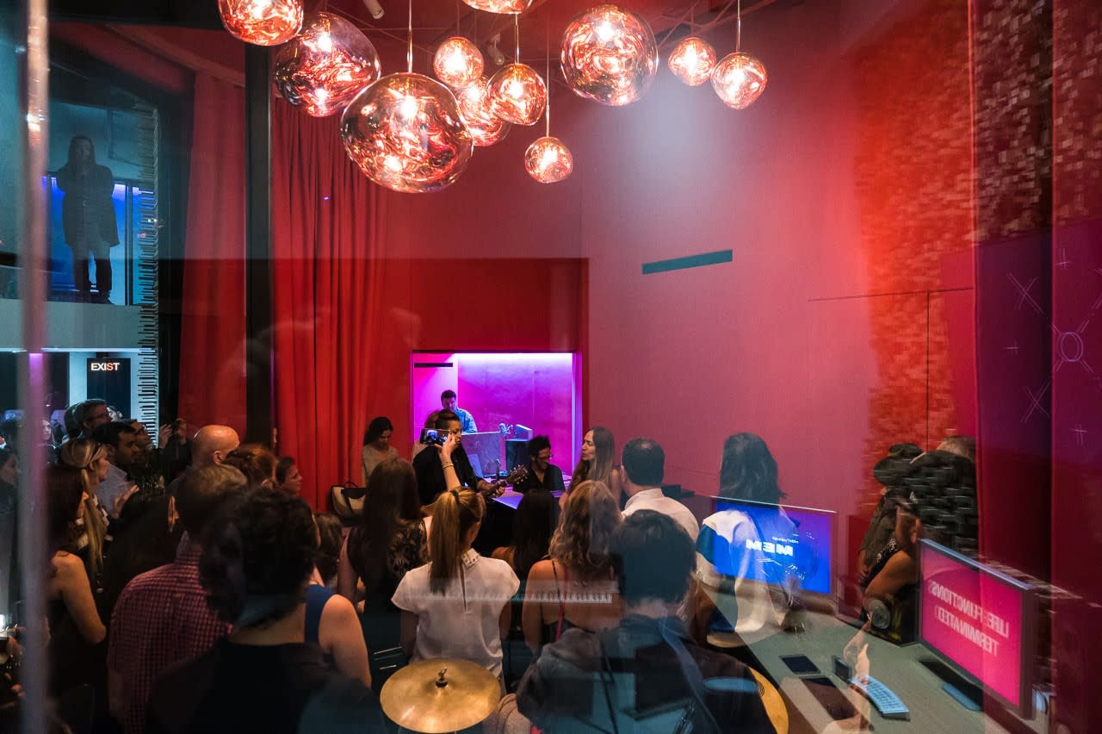 A crowded room features a live musical performance with musicians and an audience illuminated by decorative pendant lights.