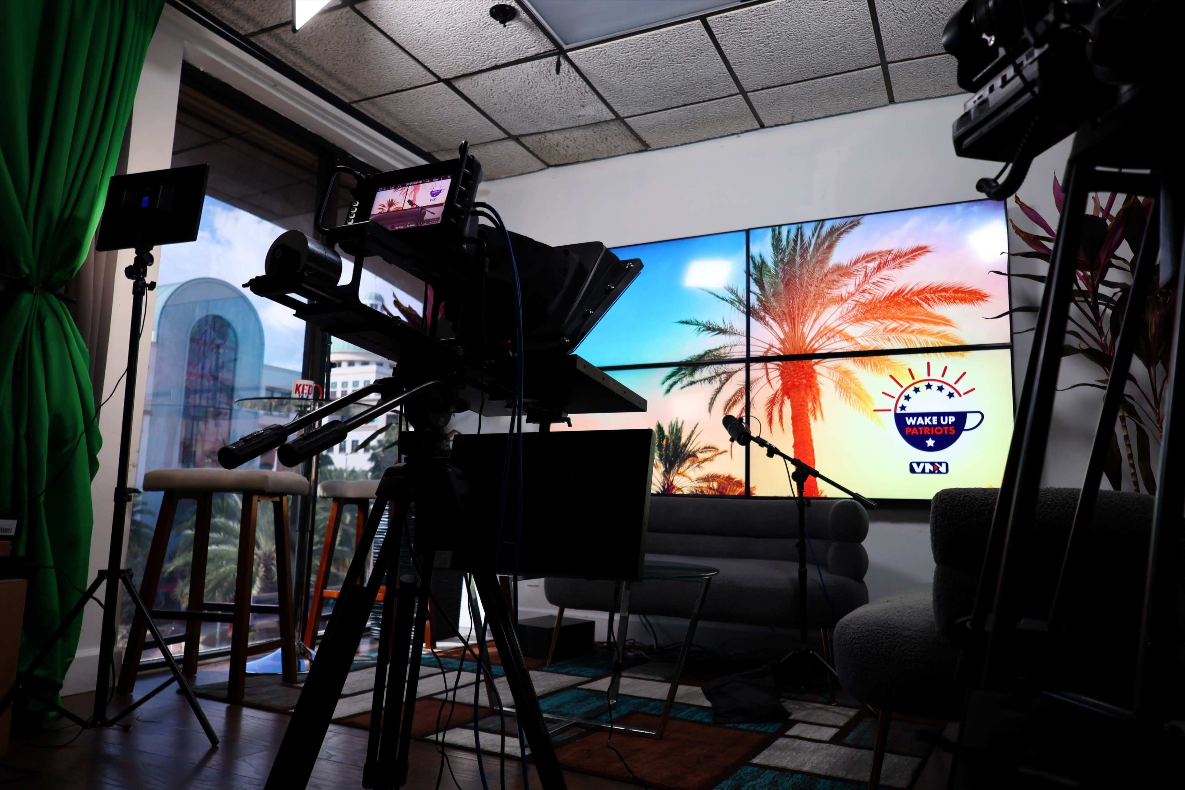 A television studio setup with cameras, a microphone, and a large screen displaying palm trees against a colorful backdrop.