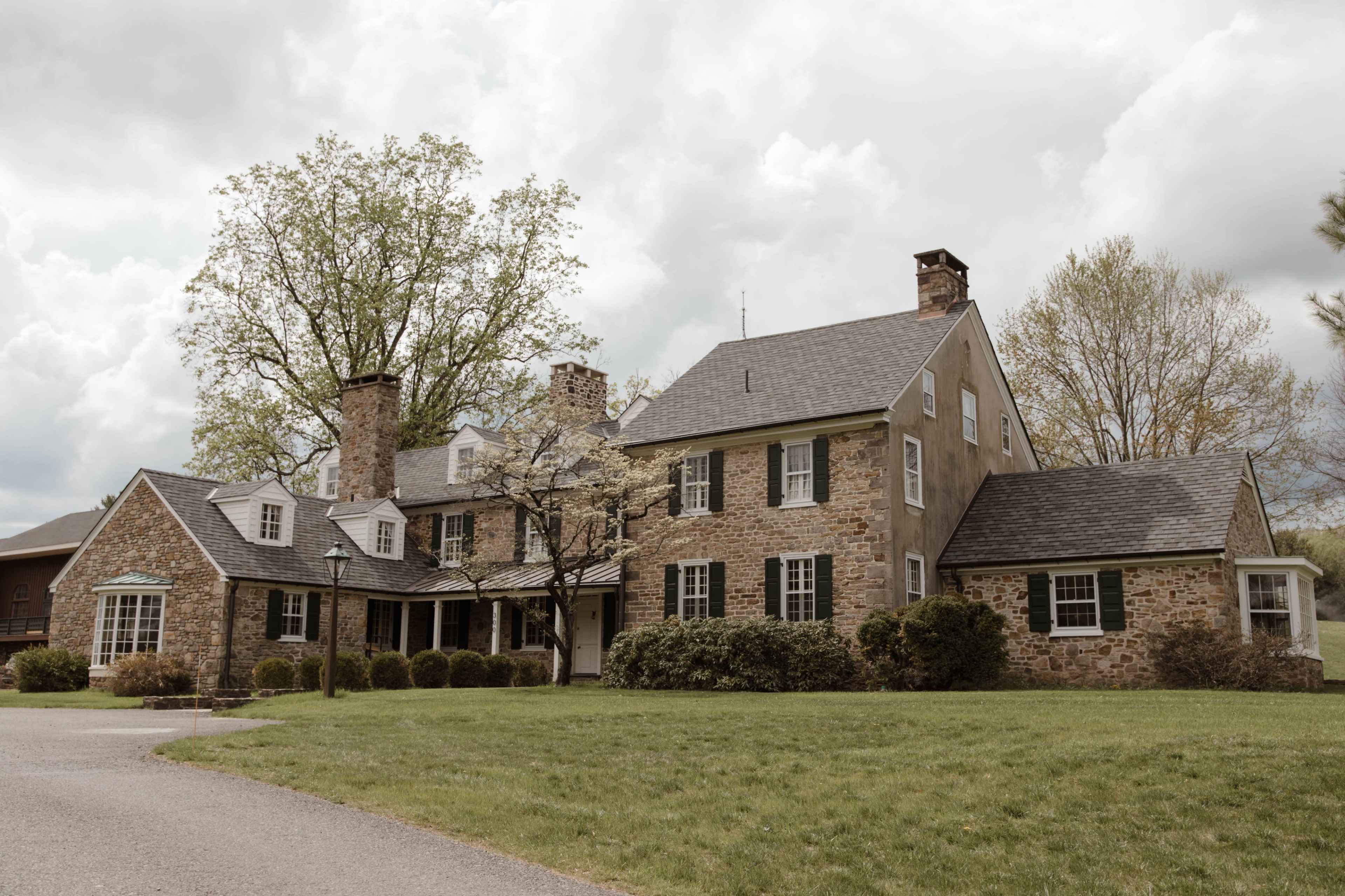 A large, stone house with multiple sections and a chimney is surrounded by trees and grassy land under a cloudy sky.