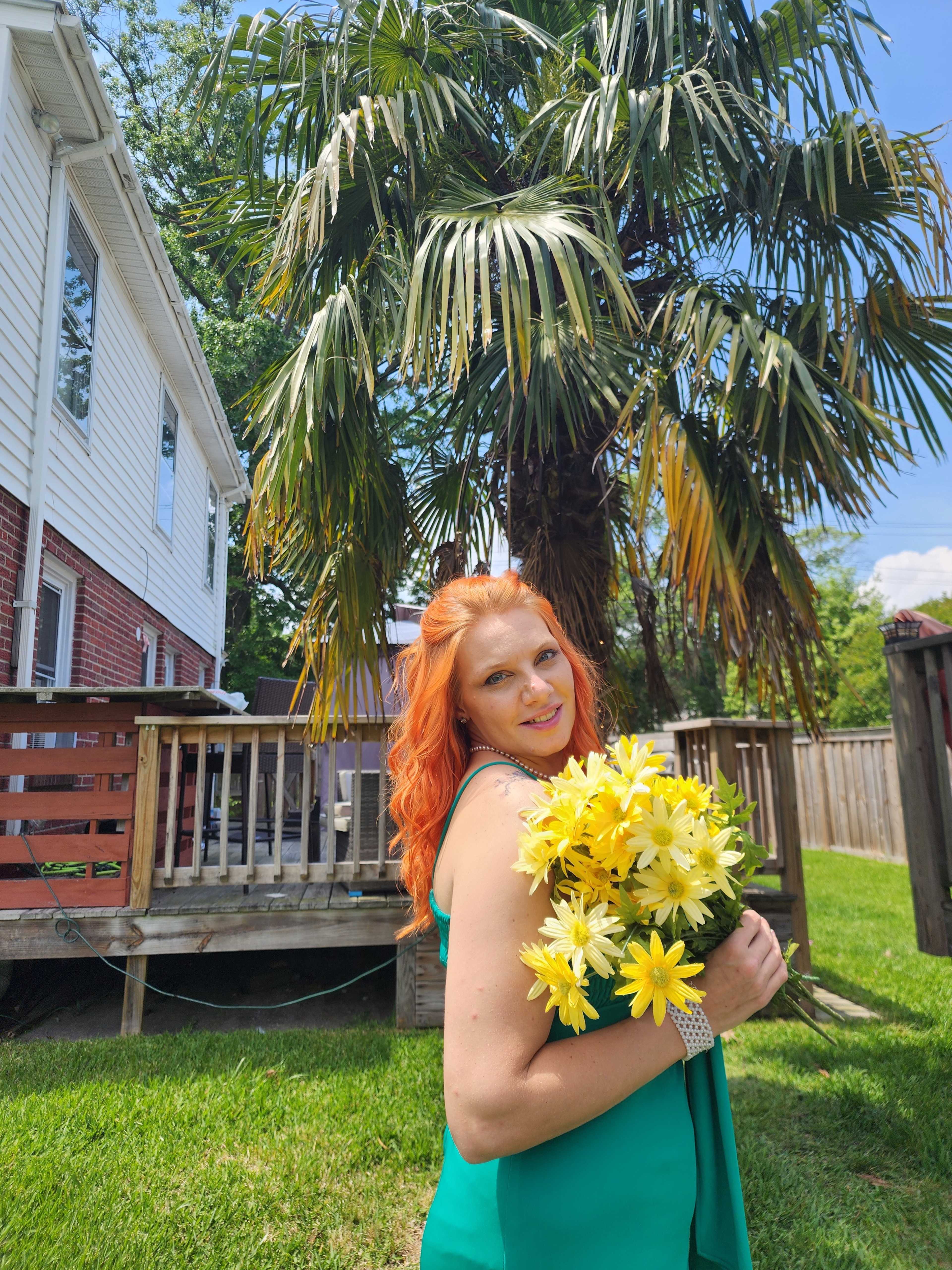 A woman with red hair holds a bouquet of yellow flowers while standing in front of a palm tree and a wooden deck.