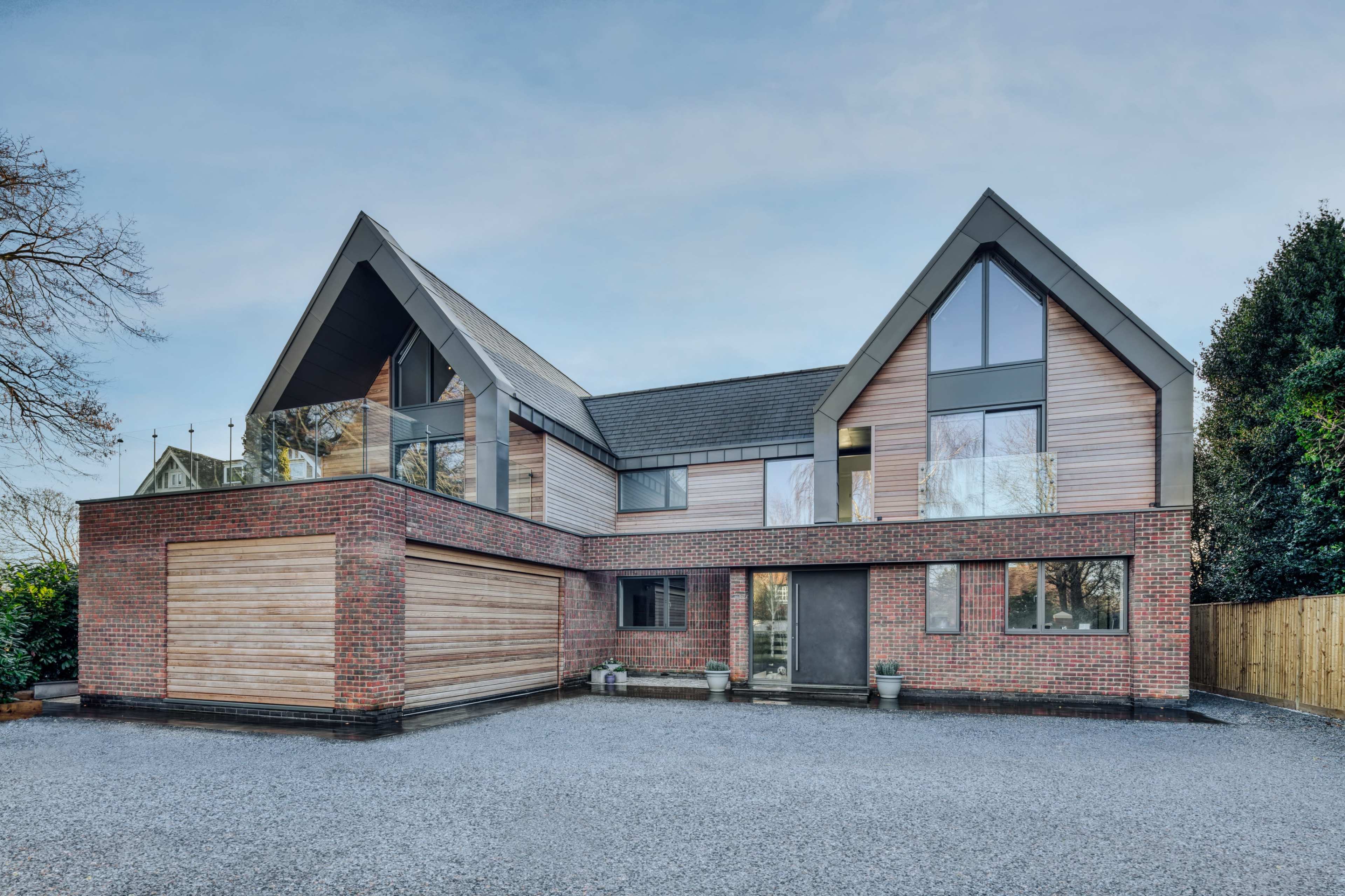 A modern two-story house features a mix of brick and wood exterior with large windows and a gravel driveway.