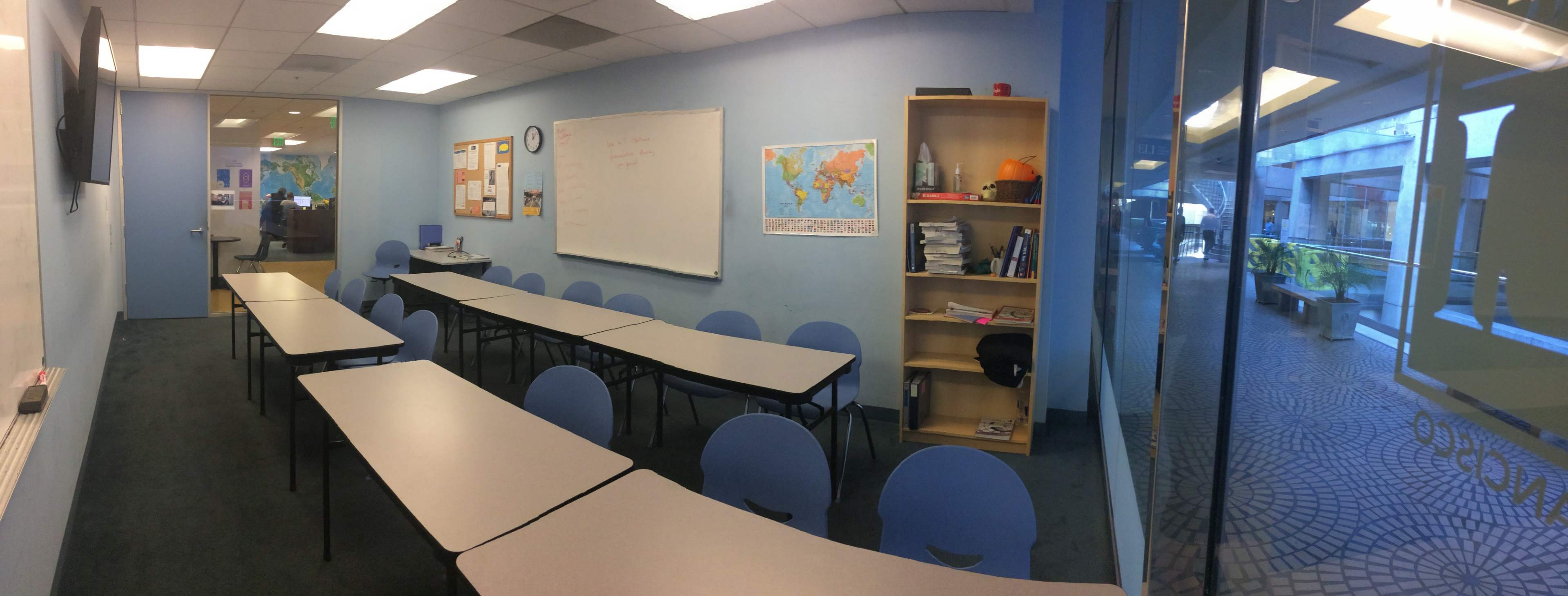 A classroom with rows of tables and chairs, a whiteboard, a map on the wall, and a bookshelf, is adjacent to a windowed area.