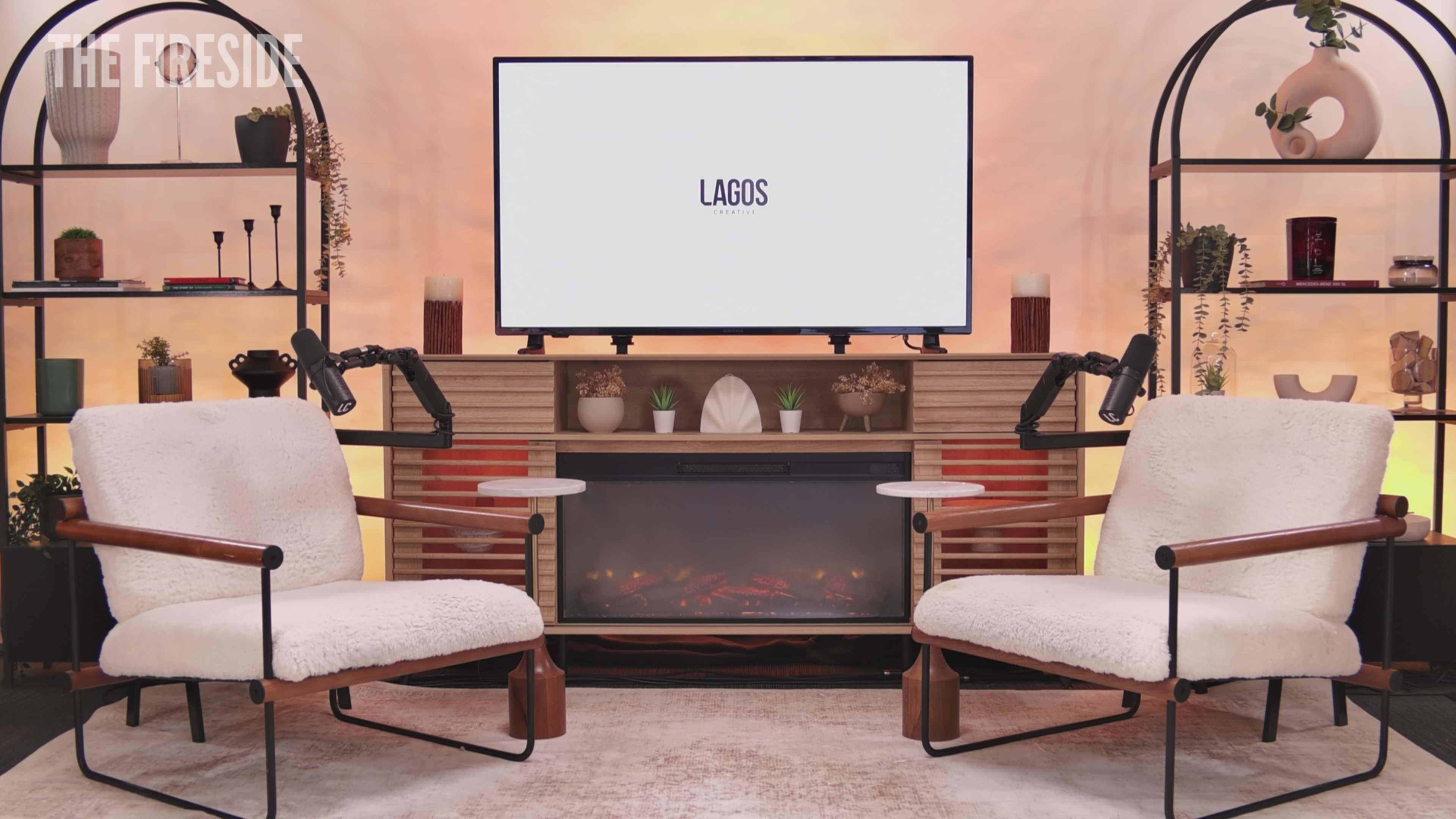 The image shows a cozy, modern studio setup with two upholstered chairs facing a wall-mounted screen displaying the word "LAGOS," flanked by decorative shelves and a faux fireplace.