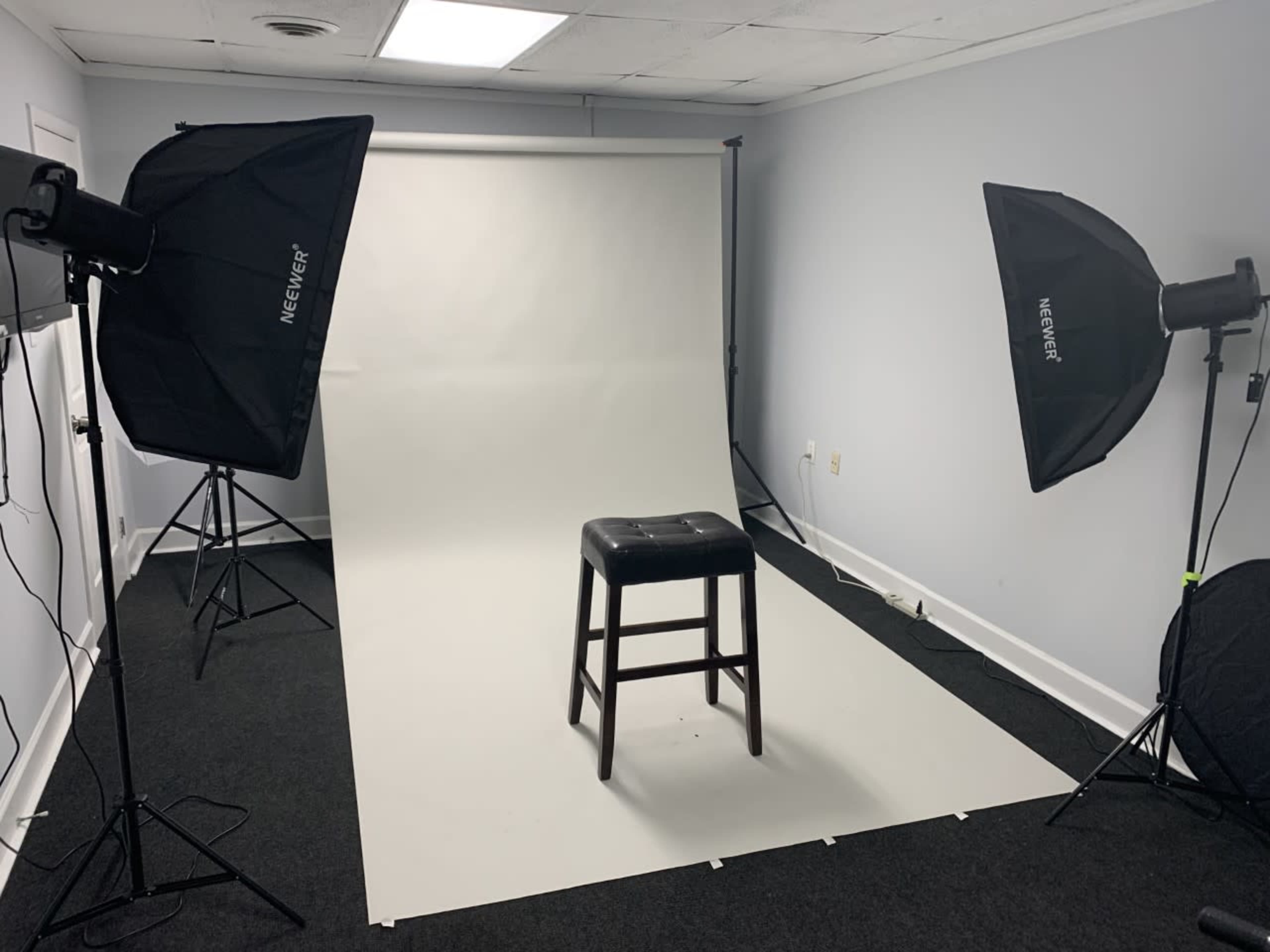 A photography studio setup with a black stool positioned on a white backdrop, illuminated by two softbox lights.