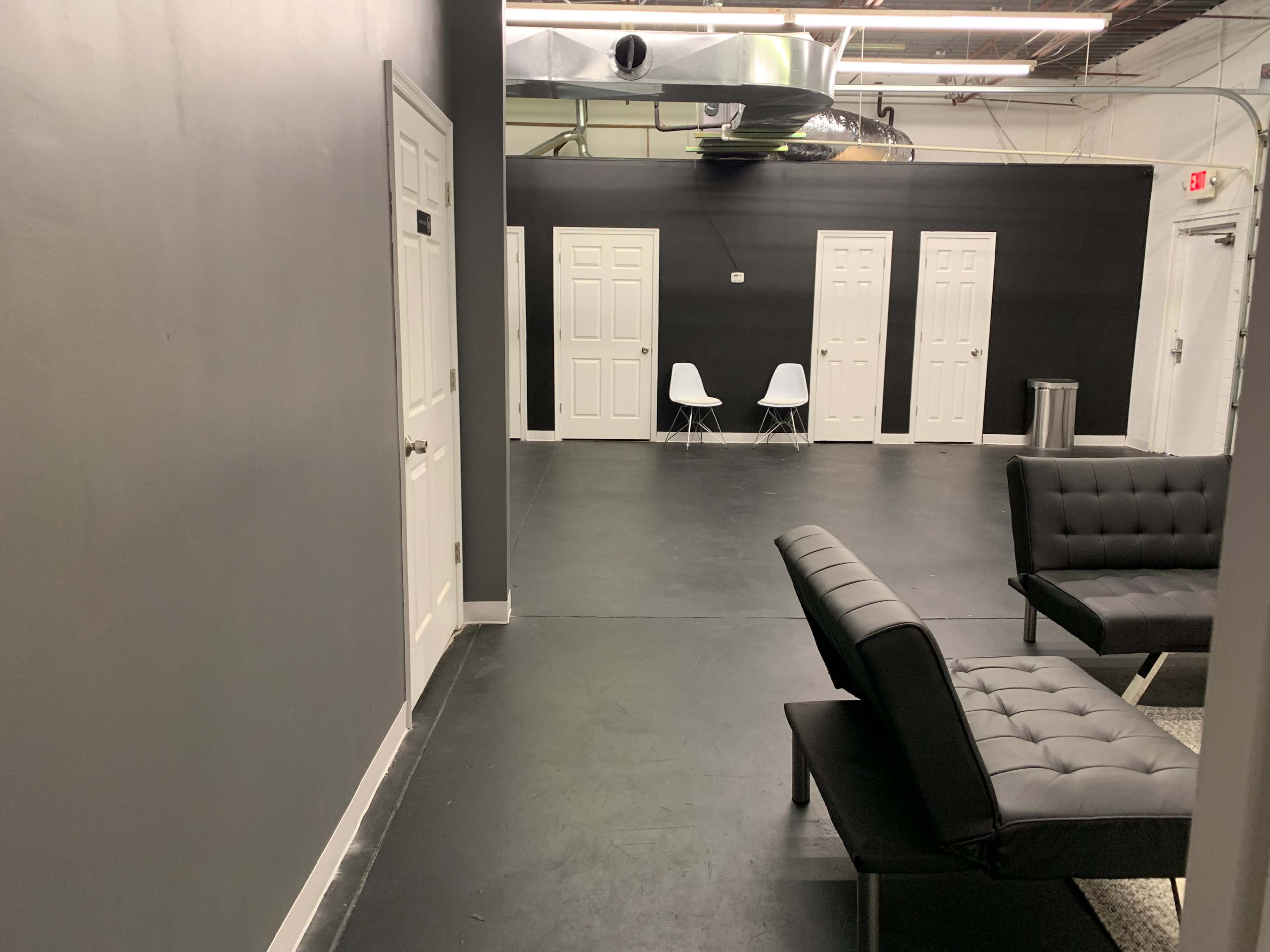 A corridor with gray walls, several closed doors, two white chairs, and black seating along the sides.