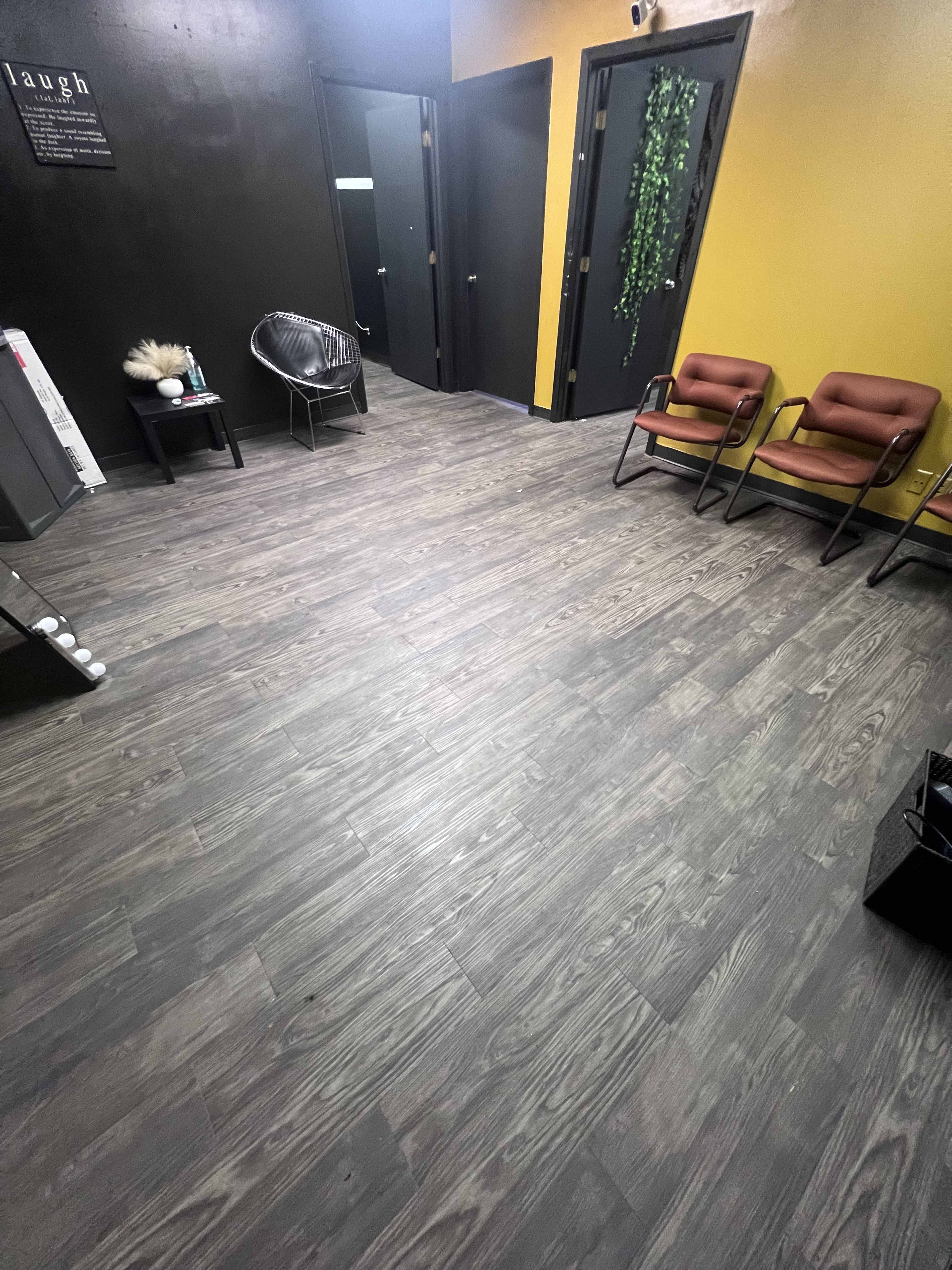 The image shows a waiting area with gray wood flooring, two brown chairs, a decorative plant on the wall, and several closed doors.