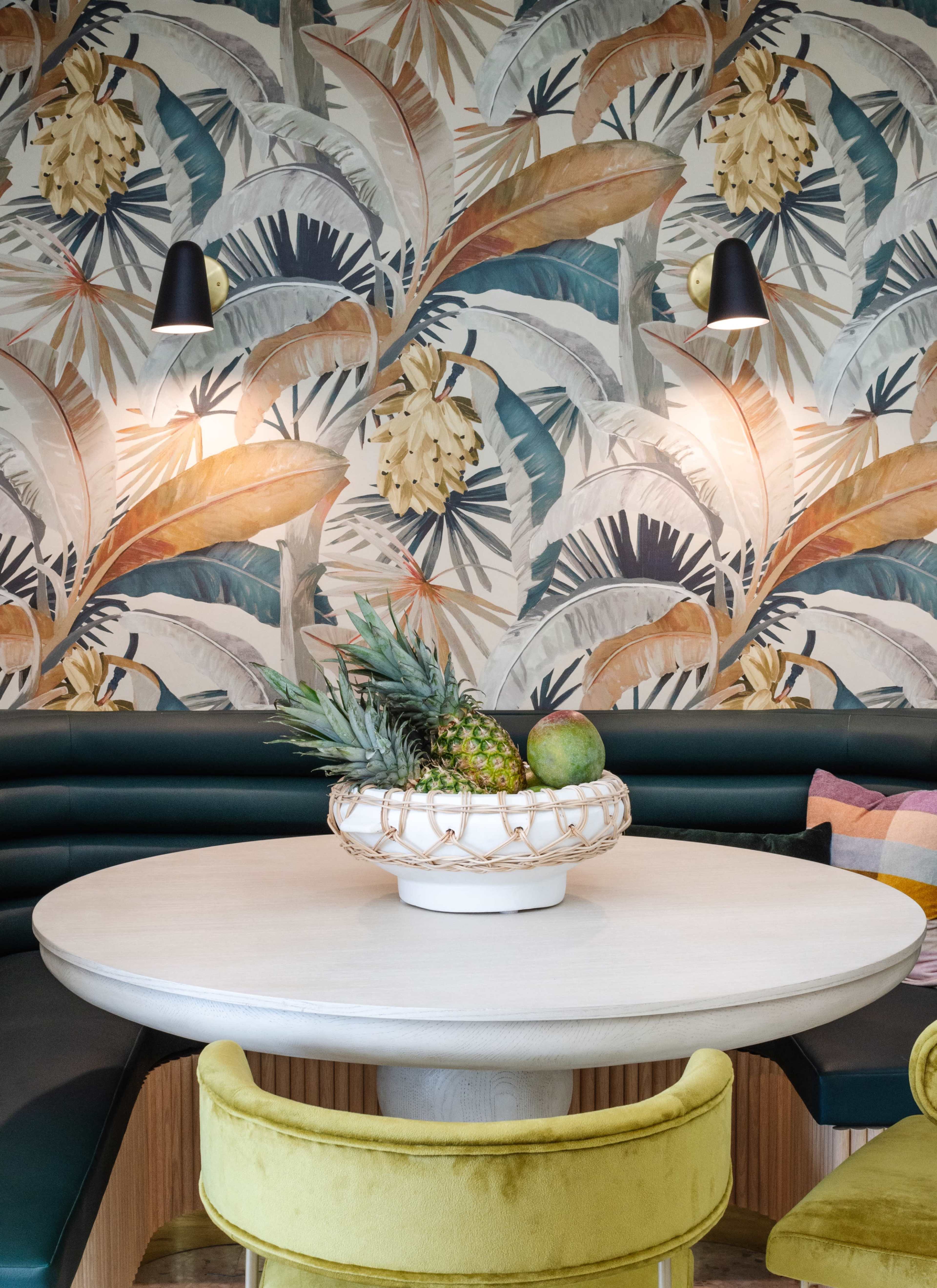 A round dining table is set in a space with a vibrant tropical leaf wallpaper, flanked by green upholstered seating and a decorative fruit bowl at the center.