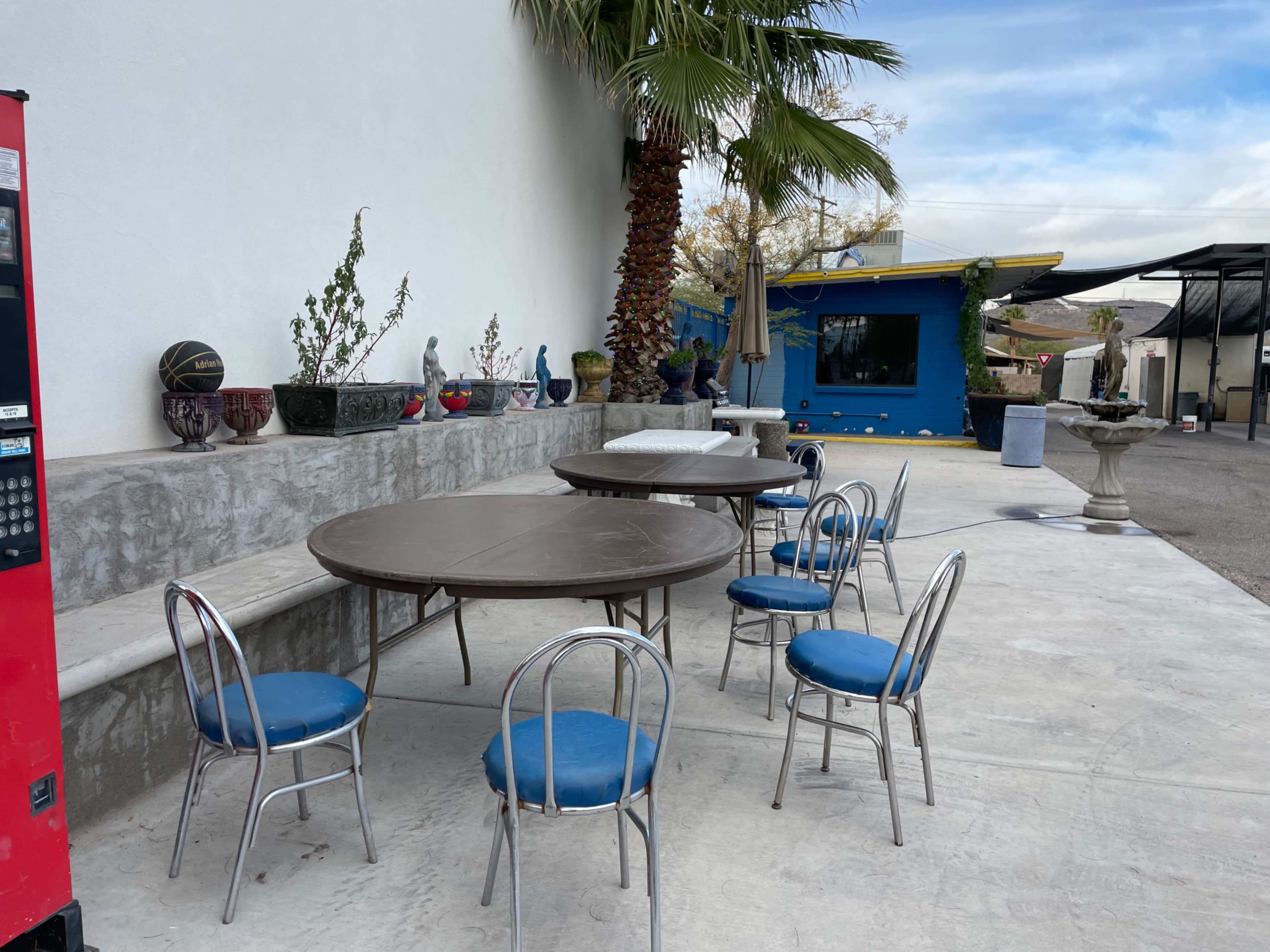 A patio area features a round table surrounded by six blue chairs, with decorative pots and a vending machine nearby.