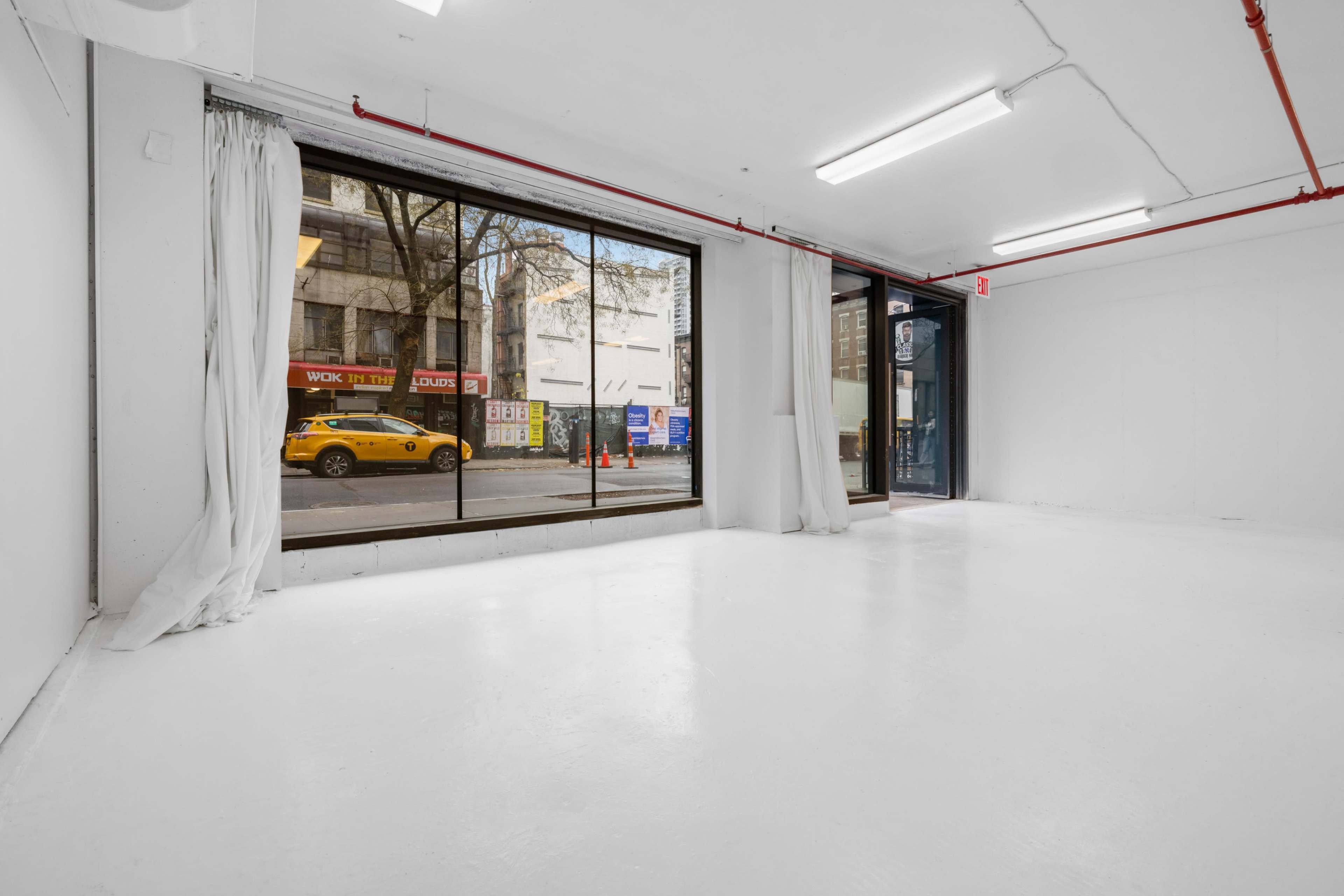 An empty, well-lit interior space with a large window facing a street, featuring a yellow taxi parked outside.