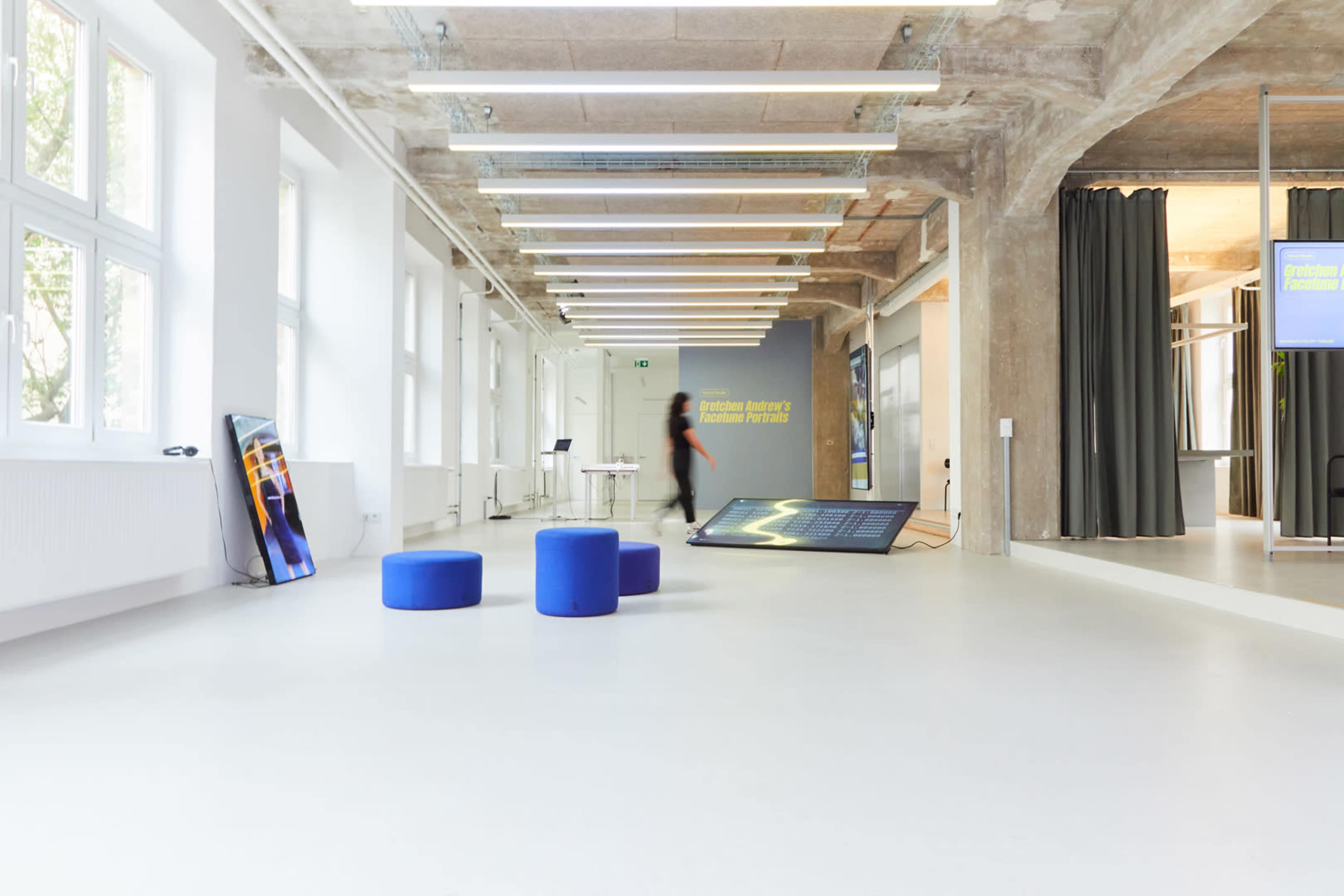 A spacious, modern interior features a white floor, multiple light fixtures, and blue seating stools, with a large digital display against a concrete wall.