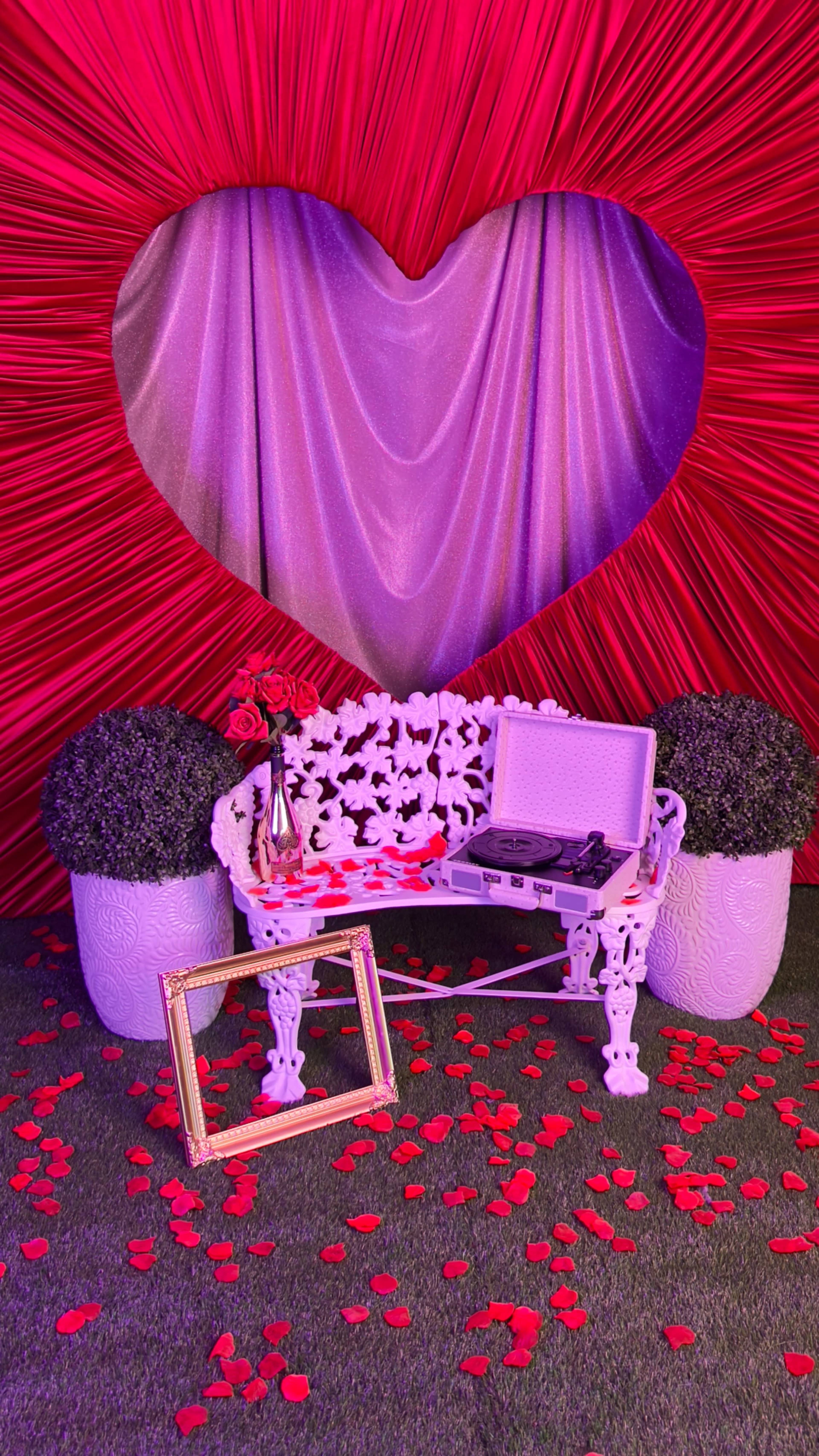 A decorative scene features a white bench surrounded by potted plants, with a heart-shaped backdrop and scattered rose petals on the ground.